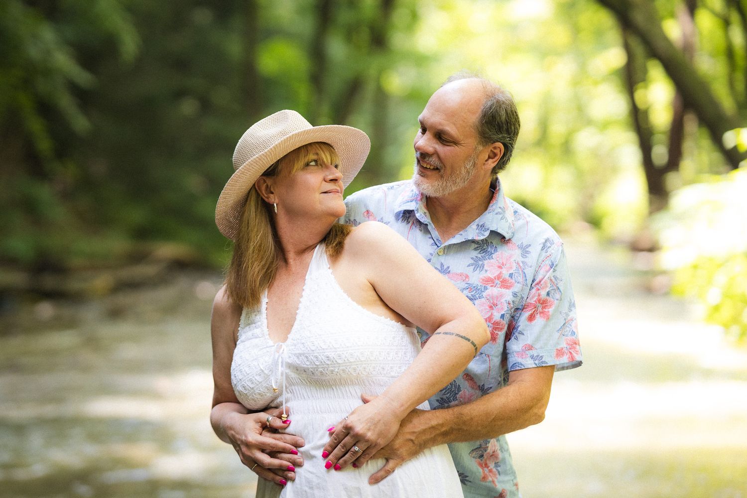 Romantic mature couple embracing outdoors in a sunlit green forest, woman wearing a straw hat.