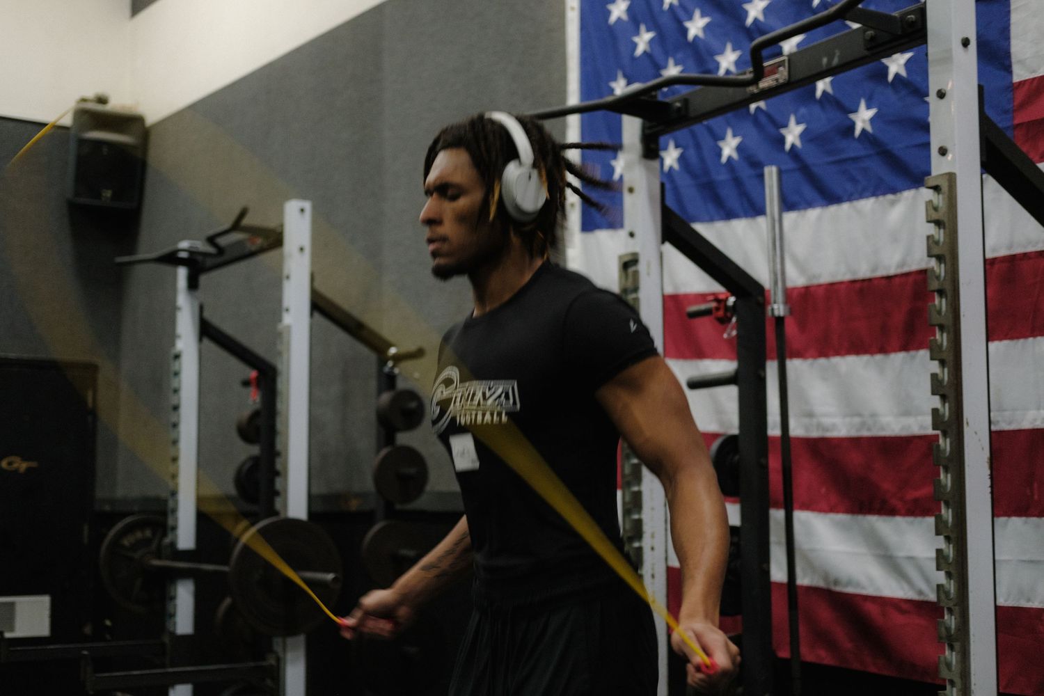 Male athlete wearing headphones doing resistance band exercises in a gym with American flag.