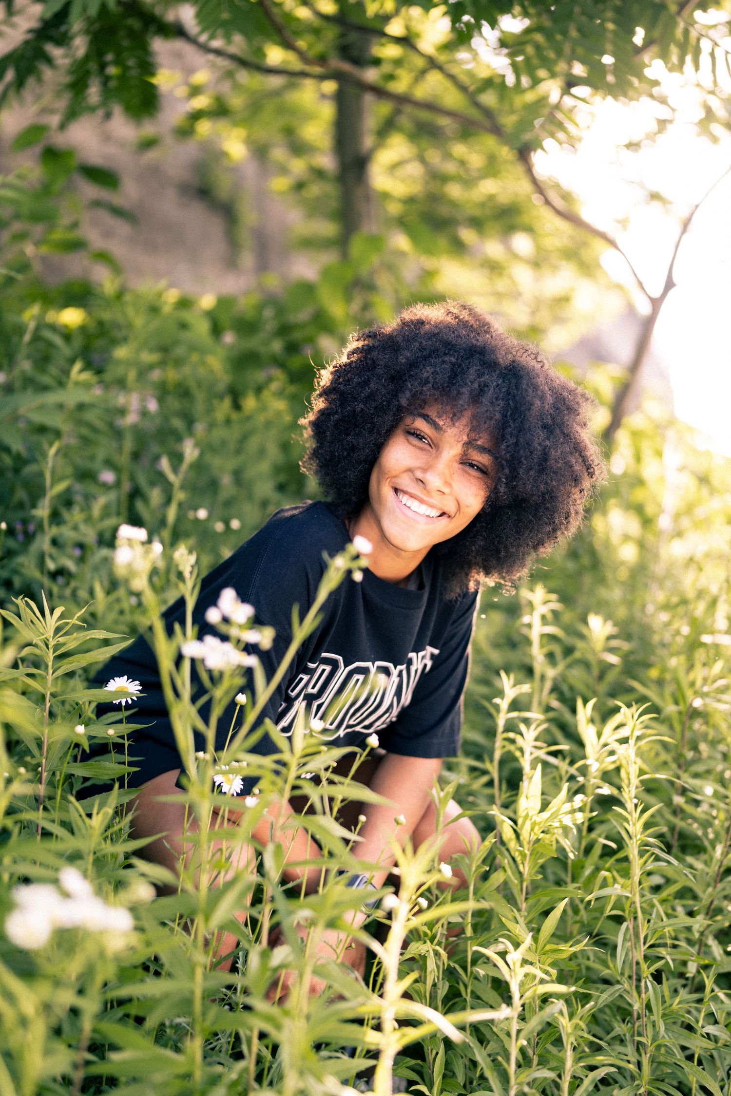 Smiling young woman with natural afro hair crouches among green plants and wildflowers.