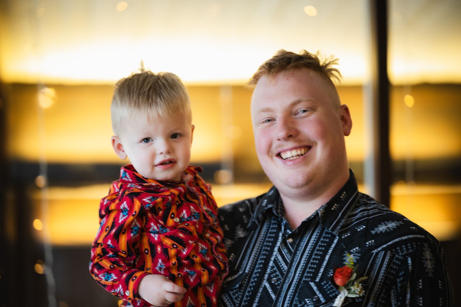 Smiling man holding a happy toddler in a red outfit at an indoor event with warm lighting.