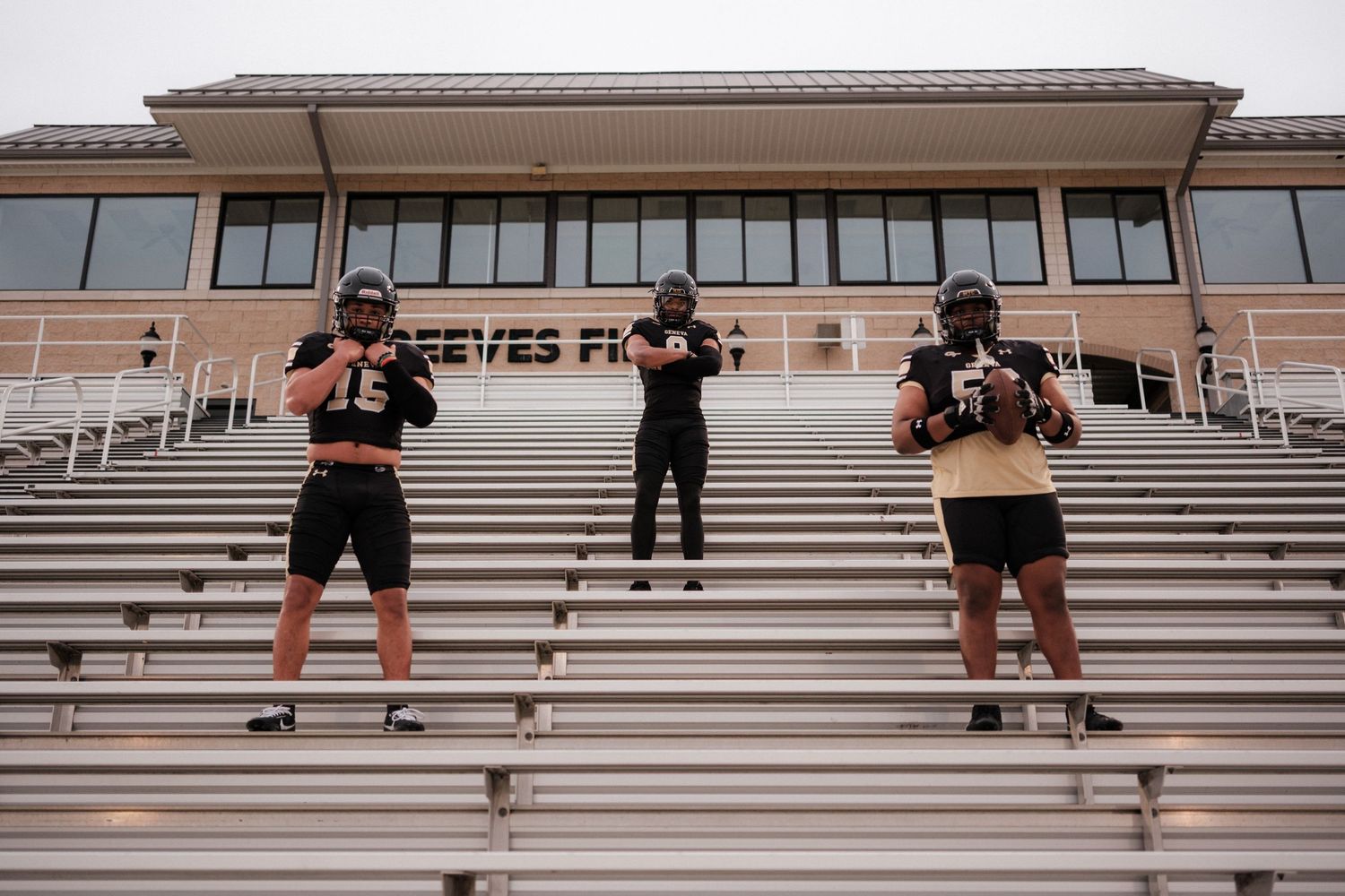 Three football players in black uniforms pose on stadium bleachers at Reeves Field.