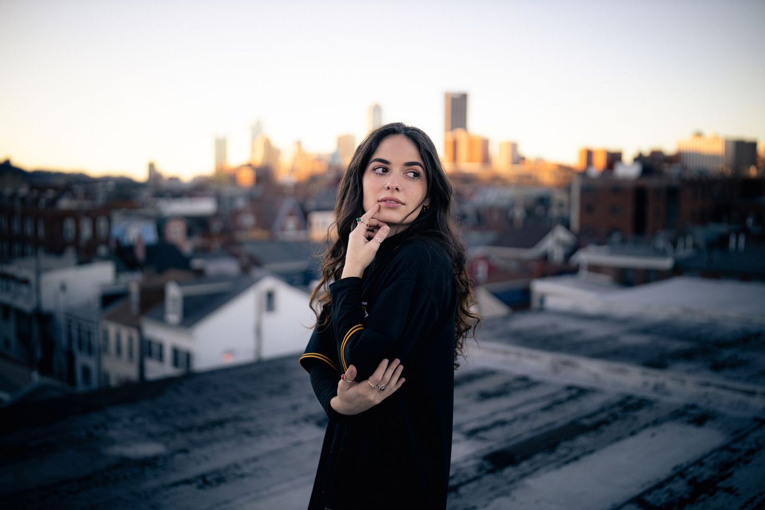 Pensive young woman with dark curly hair stands on rooftop with city skyline at dusk.