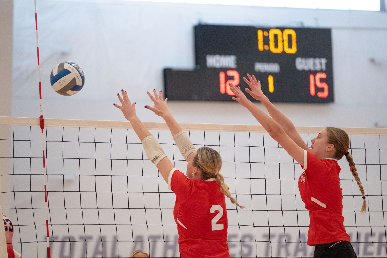 Two volleyball players in red jerseys block at the net with scoreboard showing 12-15.