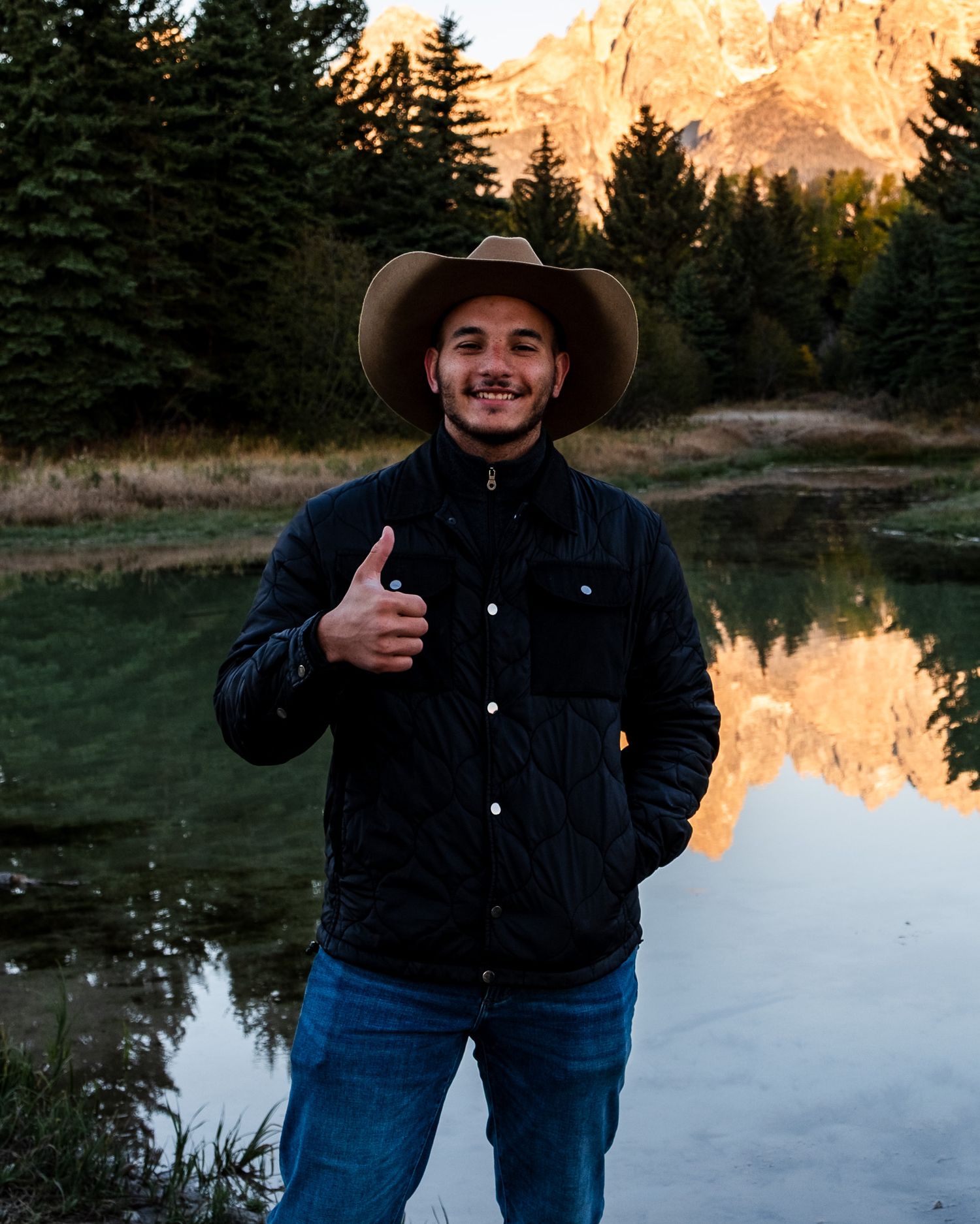 Smiling man in cowboy hat giving thumbs up by mountain lake with forest reflection at sunset.