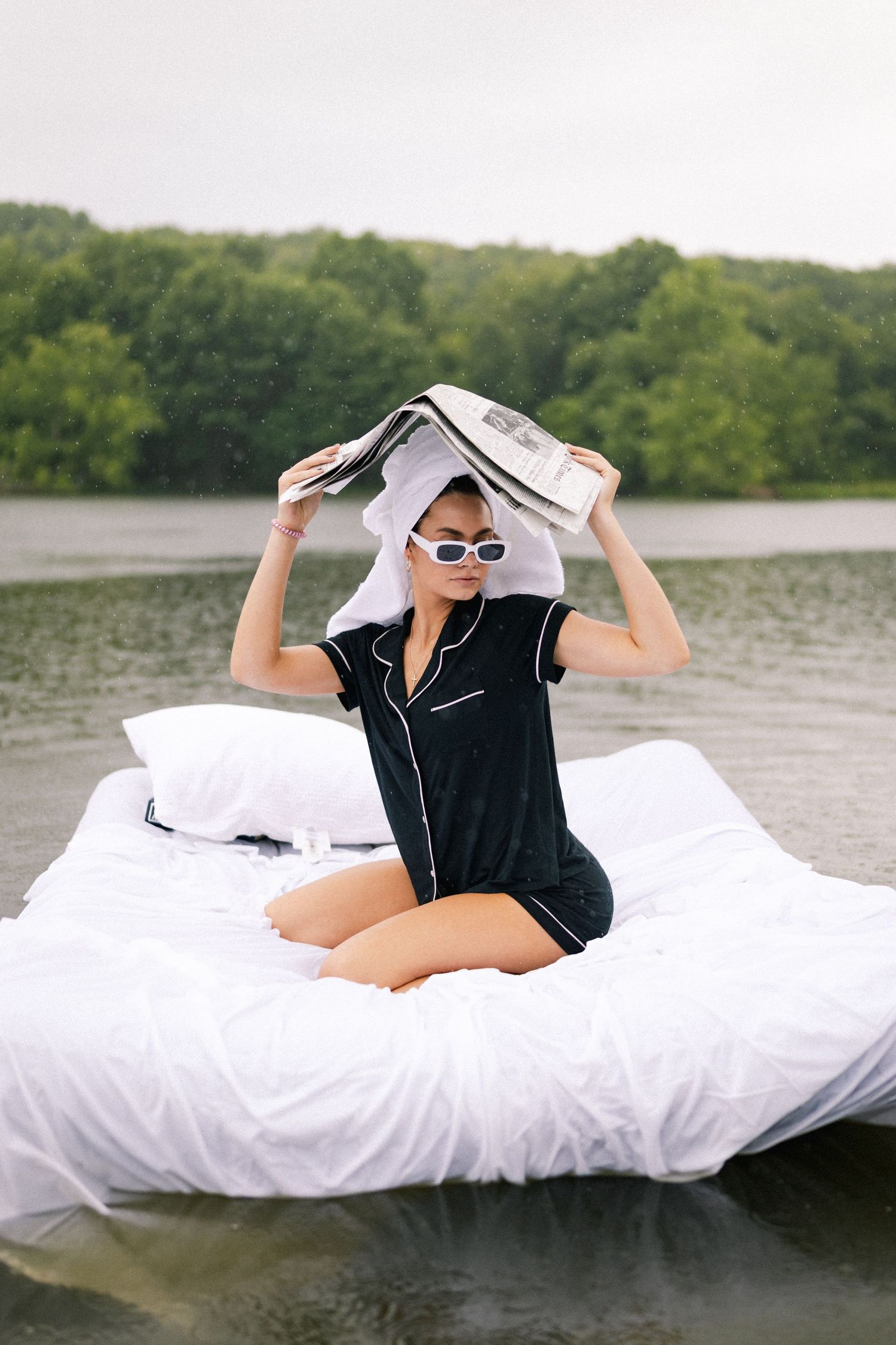 Woman in black pajamas holds newspaper over head while sitting on floating bed on lake.