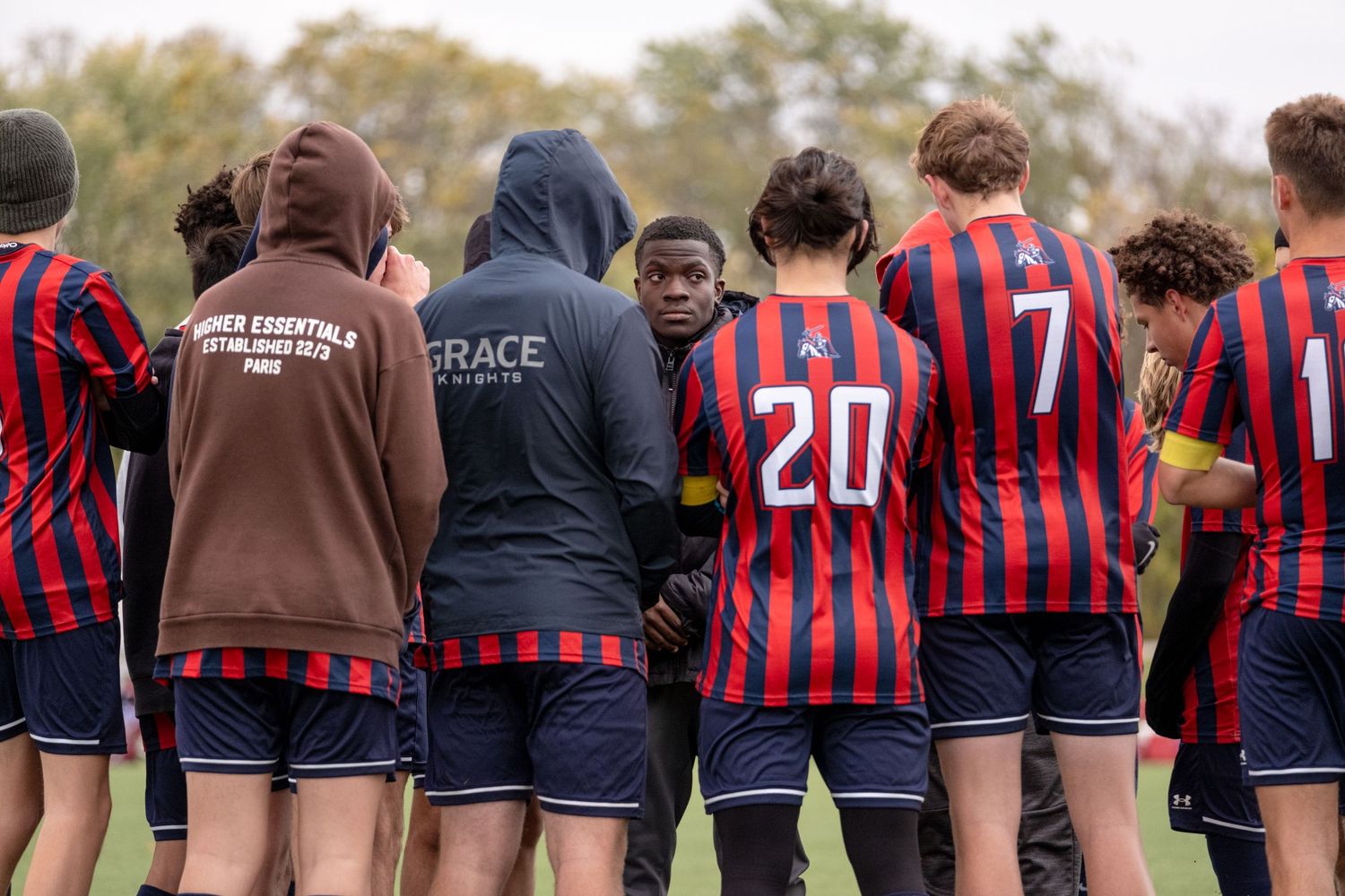 Youth soccer team huddles with coach in red and blue striped uniforms on a fall day.