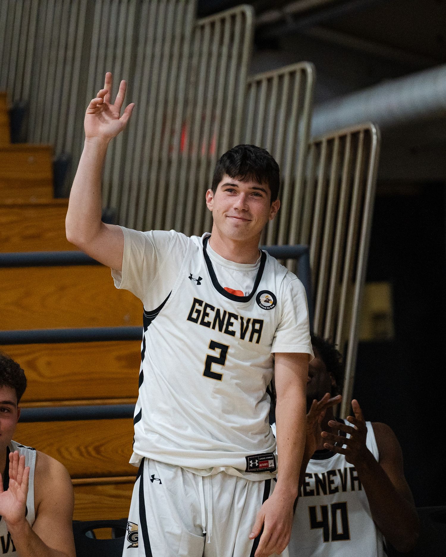 Geneva College basketball player number 2 in white uniform celebrates with three-point gesture.