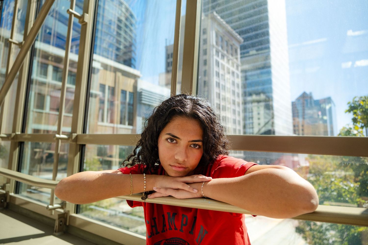 Woman in red shirt leans on railing with city skyline visible through windows.