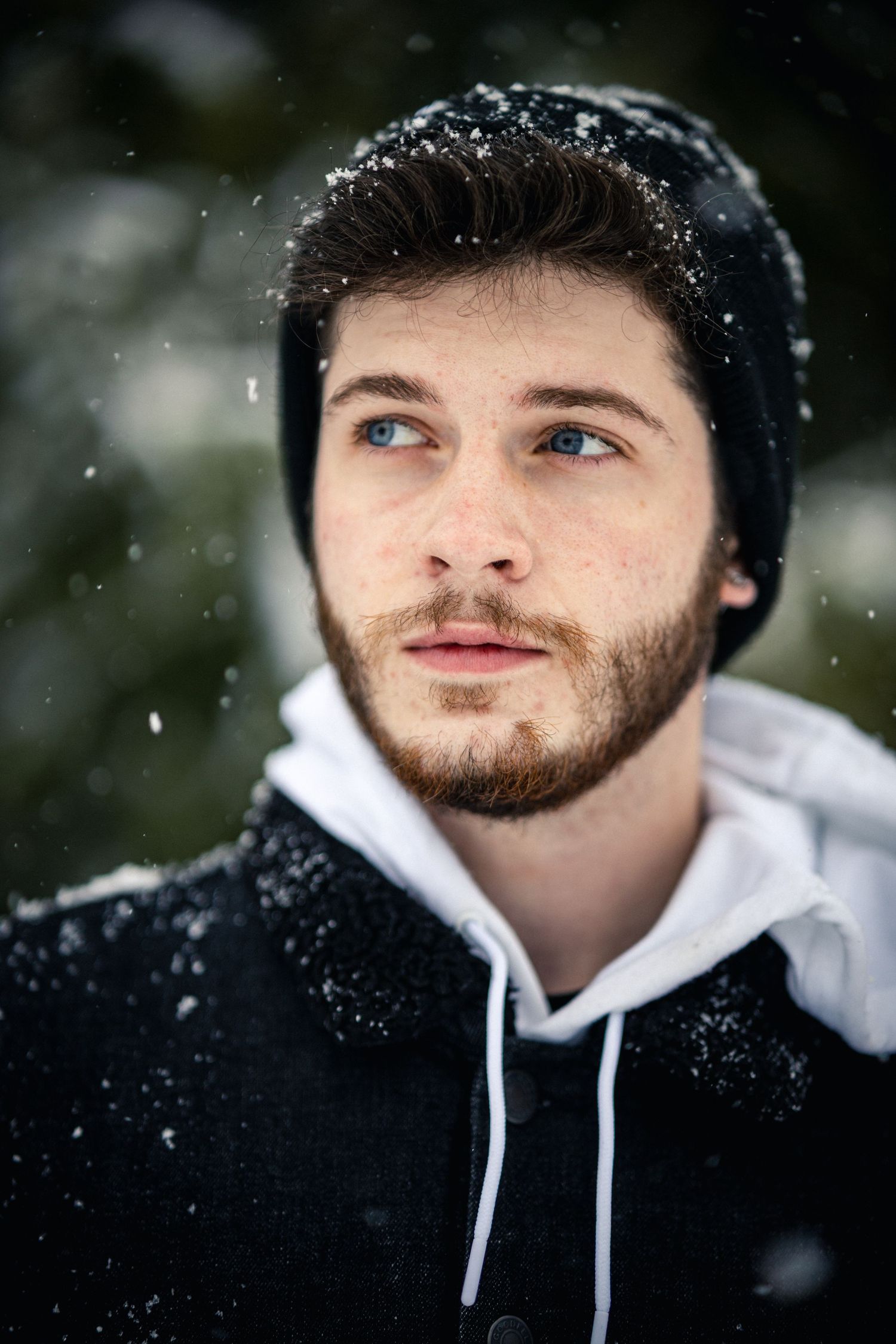 Young man with blue eyes and beard wearing a black hoodie standing outdoors in falling snow.
