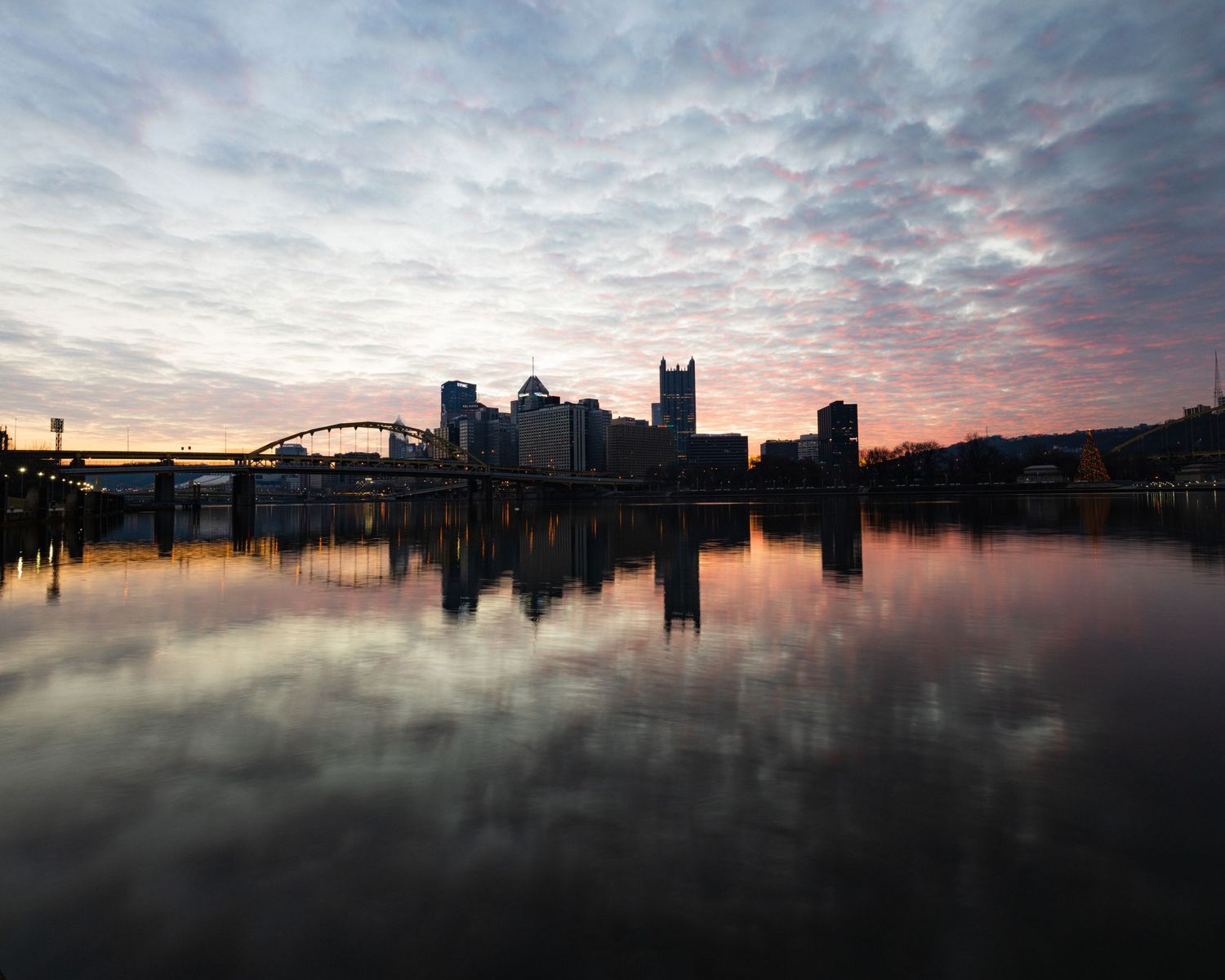 Pittsburgh skyline mirrored in still river at dusk with colorful clouds and bridge silhouette.
