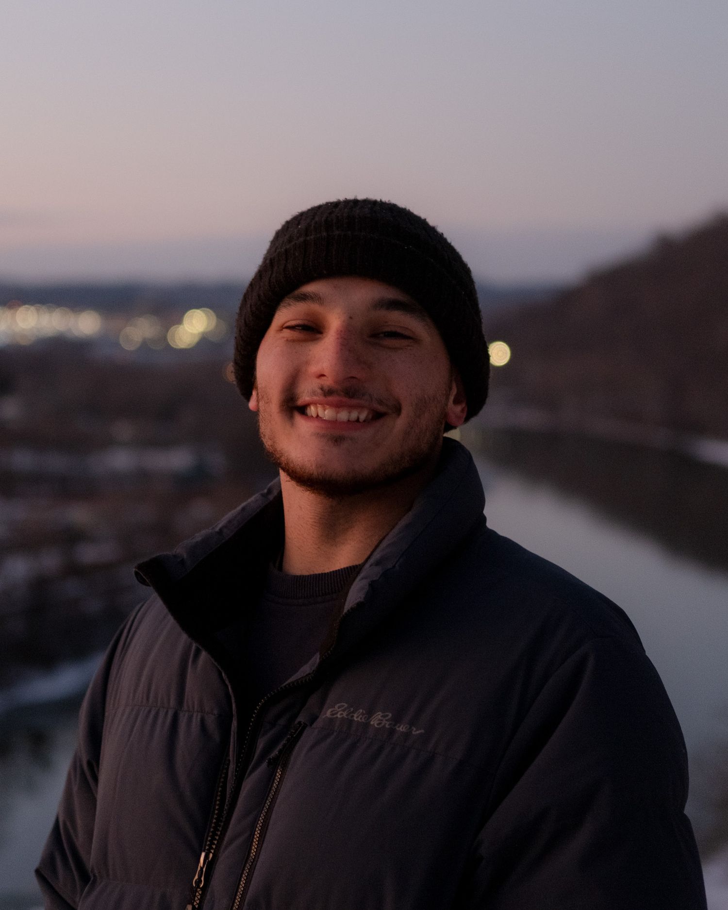 Smiling young man in black beanie and puffer jacket posing outdoors at dusk near a river.