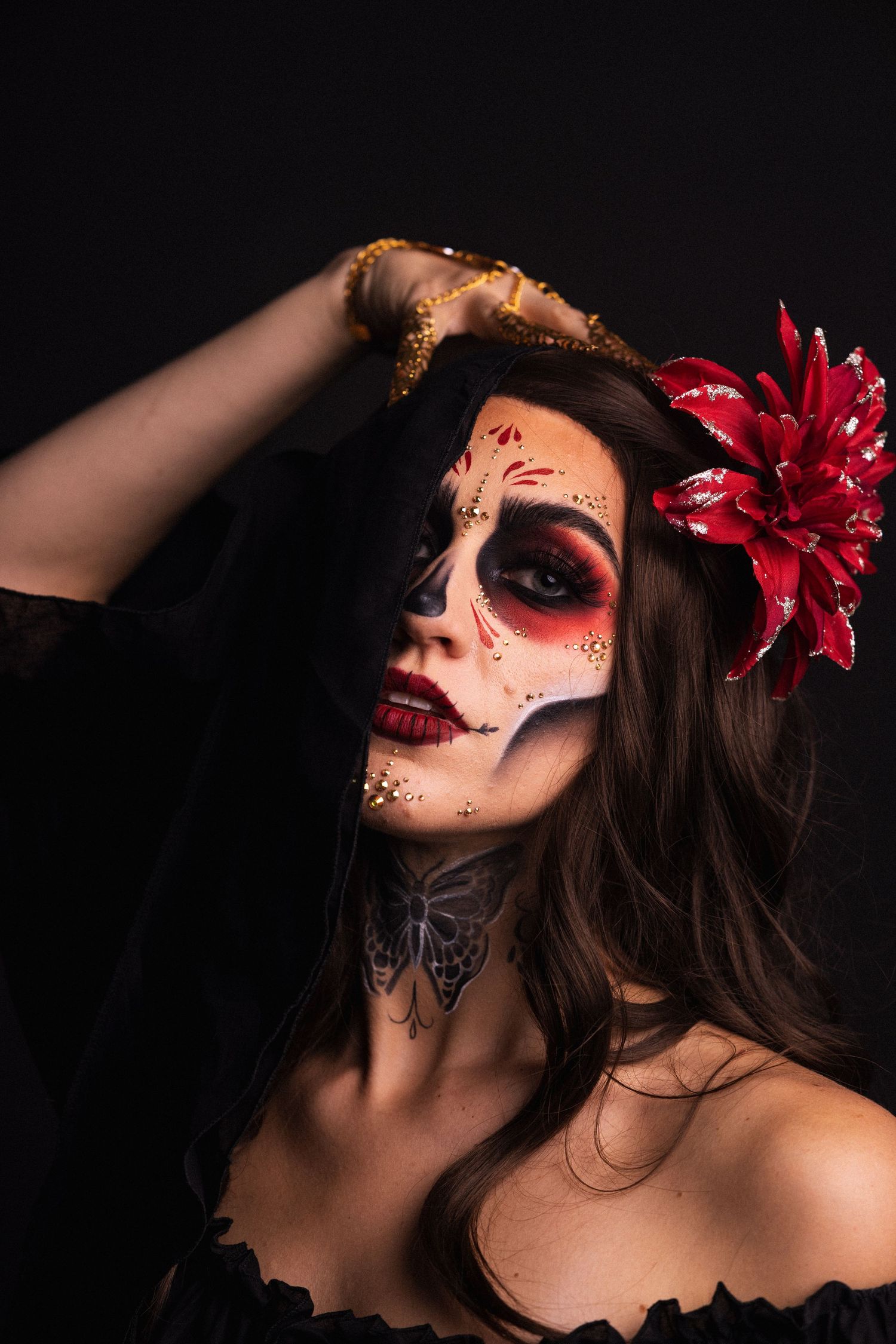 Woman with sugar skull makeup and red flower in hair poses dramatically against dark background.