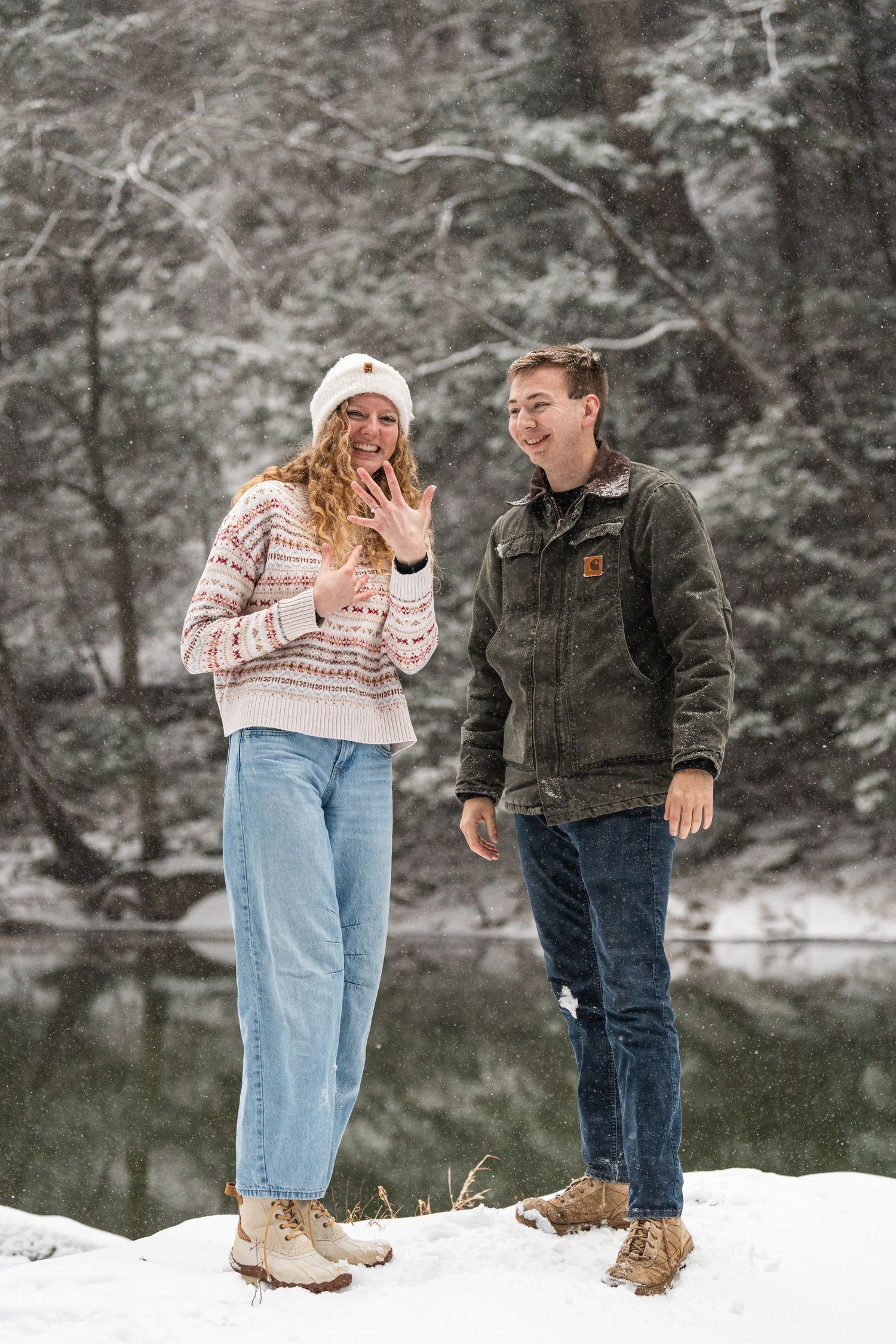 Newly engaged couple smiles and shows off engagement ring in a snowy outdoor winter setting.