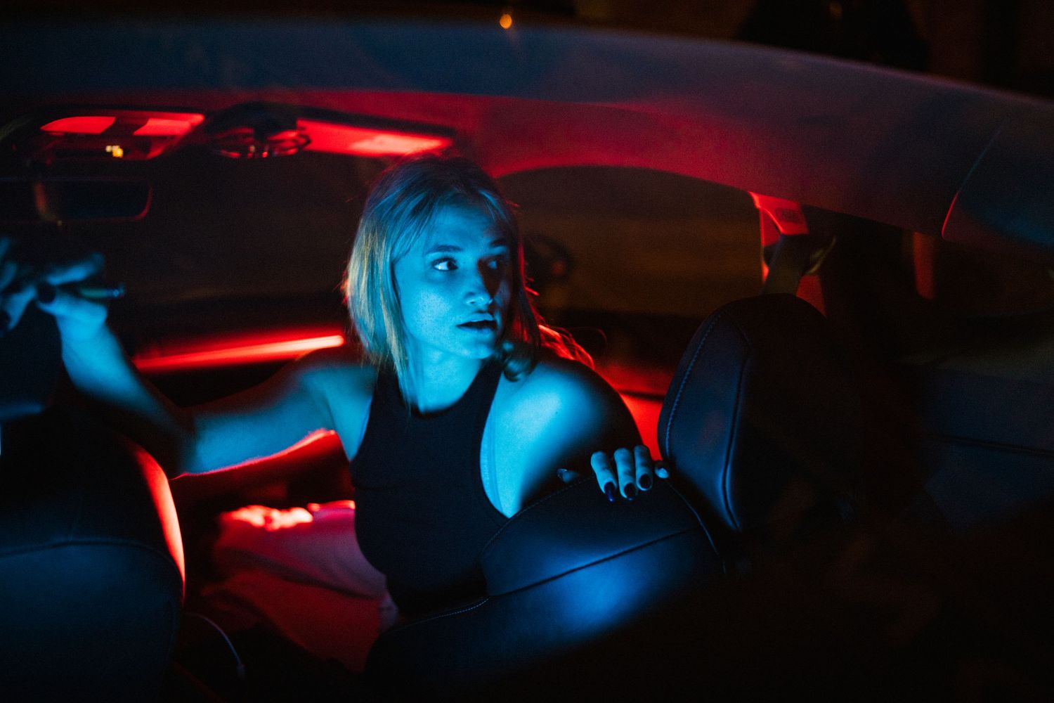 Blonde woman sits in car interior illuminated by dramatic red and blue neon lighting at night.