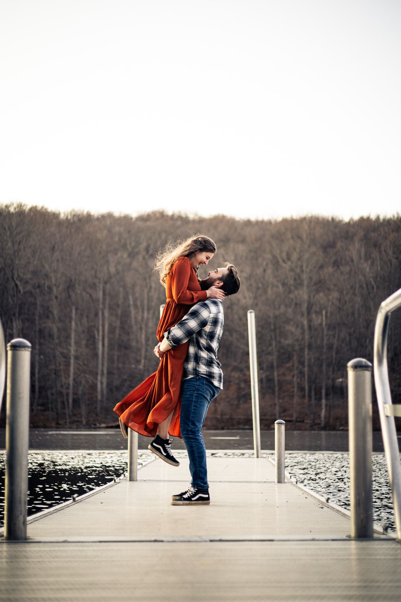 Couple embracing on lakeside dock at sunset, woman in flowing rust dress lifted by partner.