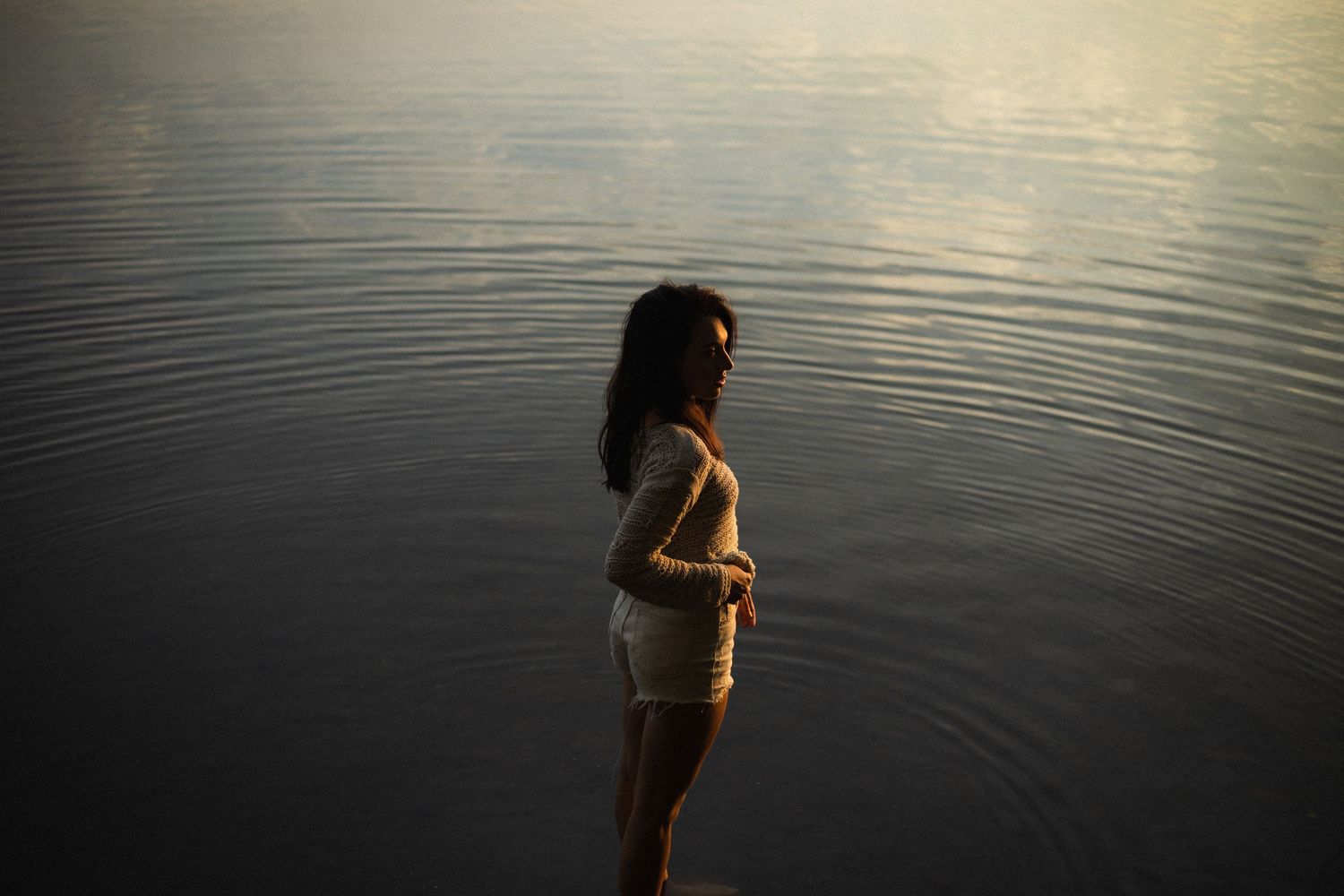 Young woman standing in shallow calm water during golden hour with soft light reflecting on the surface.