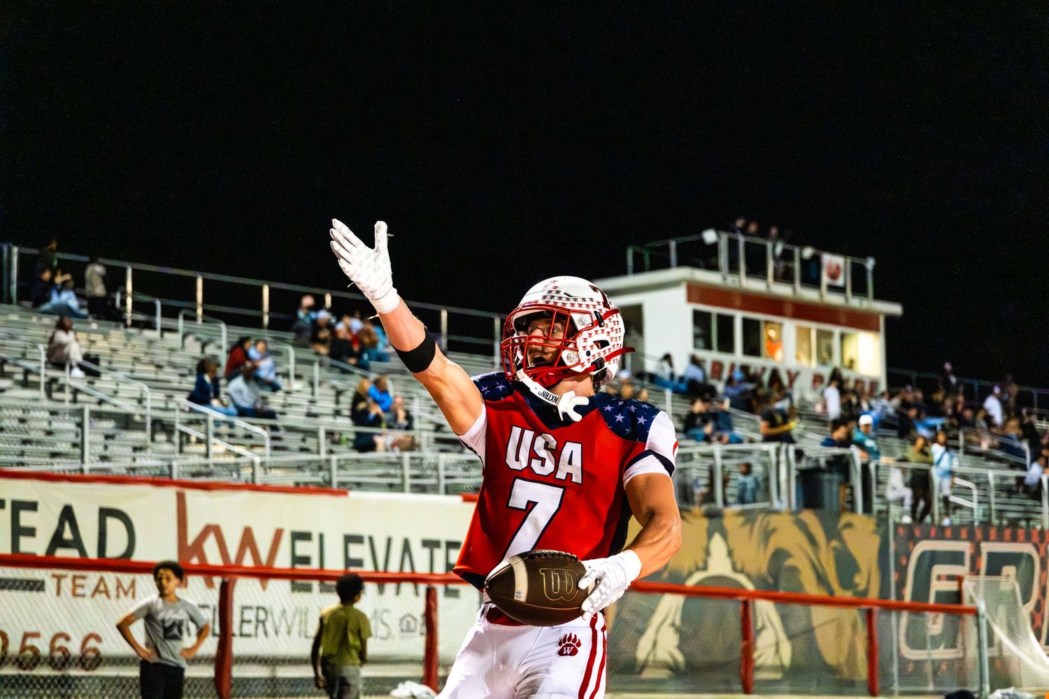 High school football player in USA jersey number 7 celebrates touchdown with one arm raised at night game.