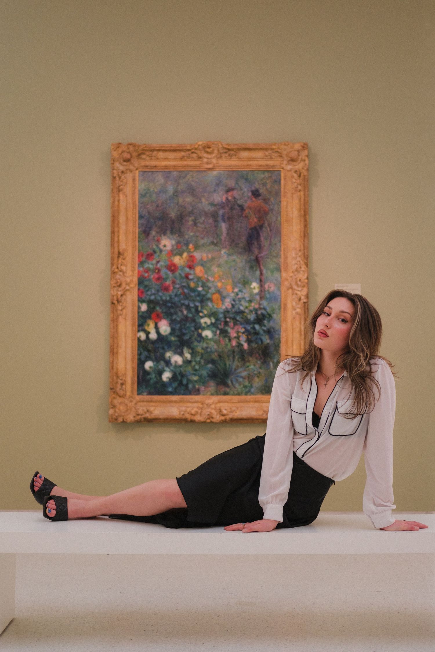 Woman in white blouse and black skirt posing on a bench in front of a framed Impressionist floral painting.
