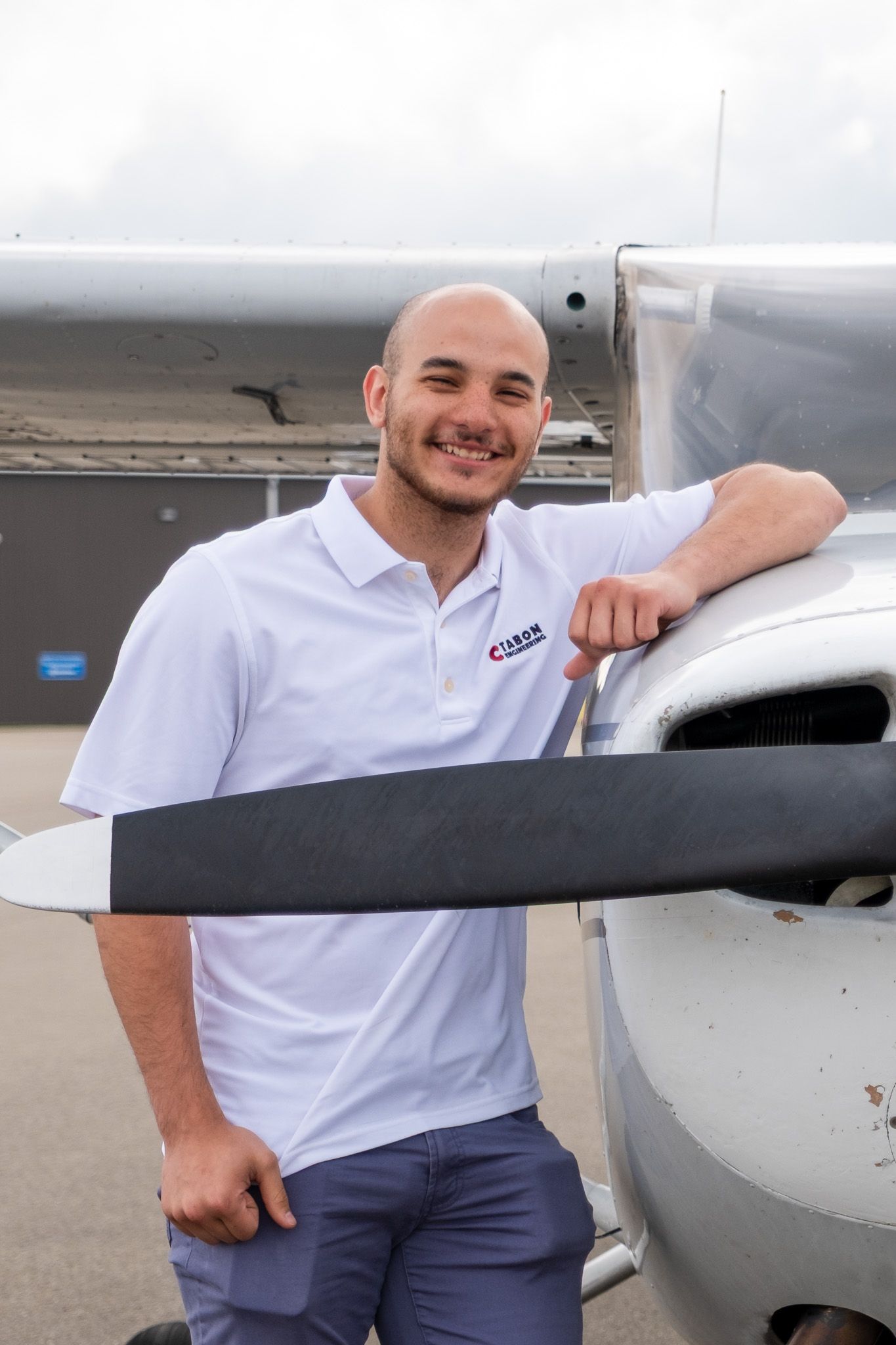 Smiling bald man in white polo shirt leaning on small aircraft propeller at airport.