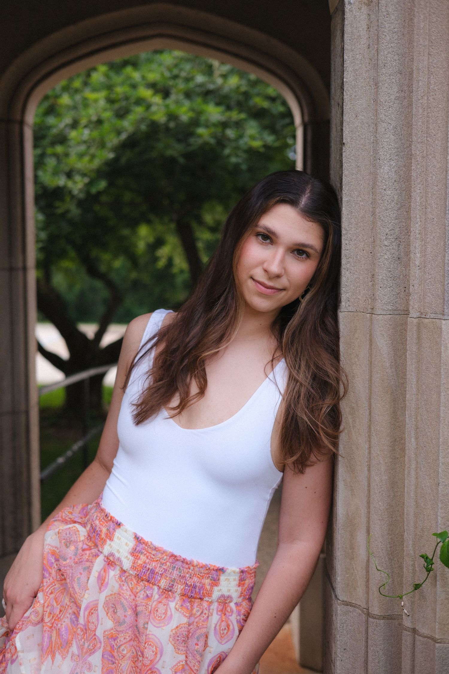 Young woman in white tank top and floral skirt leaning against a stone column in an outdoor archway.