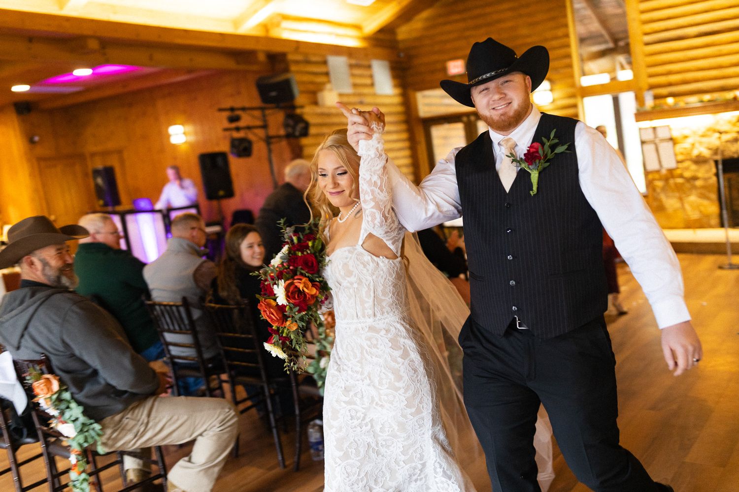 Bride and groom making a grand entrance into rustic log cabin wedding reception with bouquet raised high.