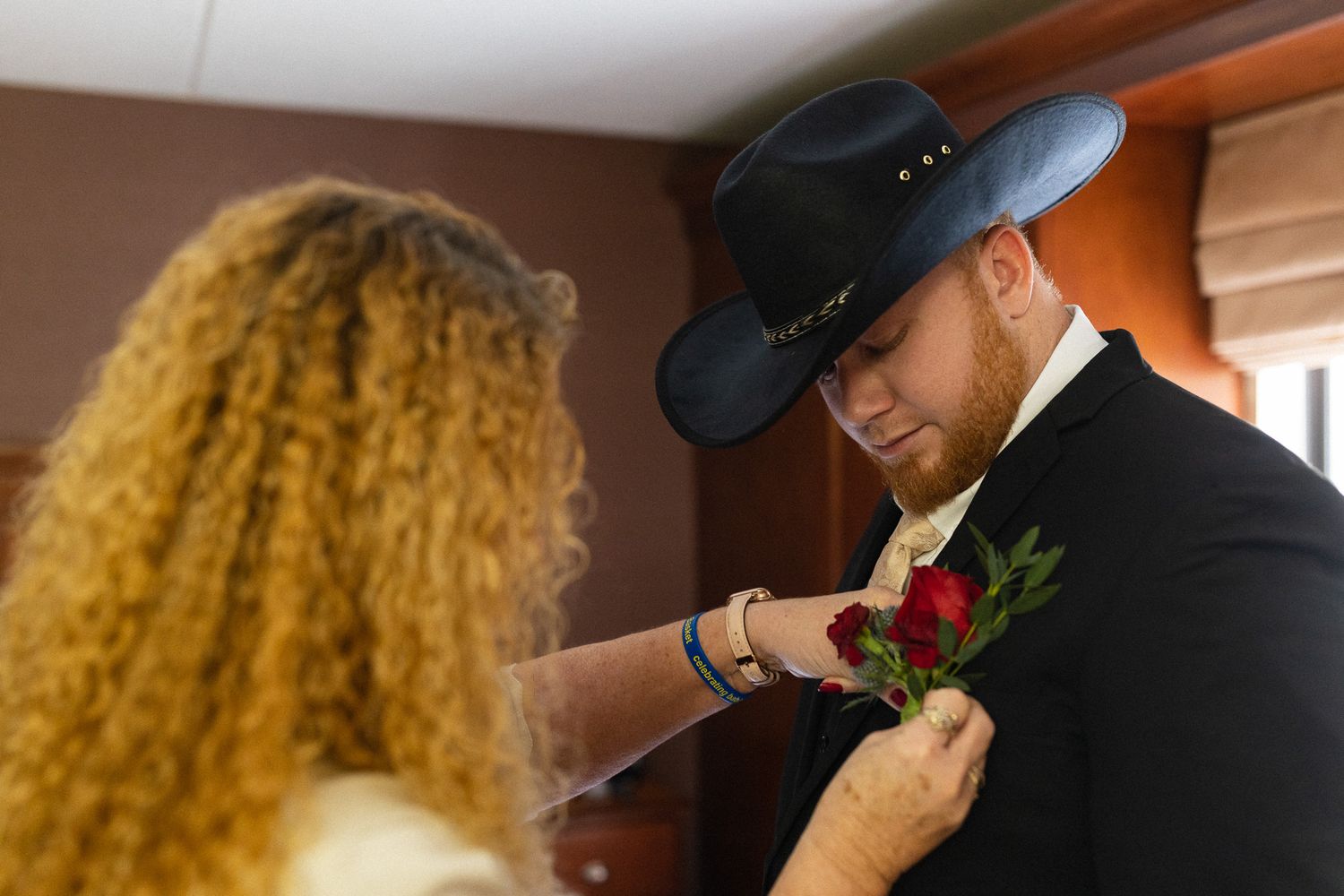 Bride pinning a red rose boutonniere on groom wearing a black cowboy hat before wedding ceremony.