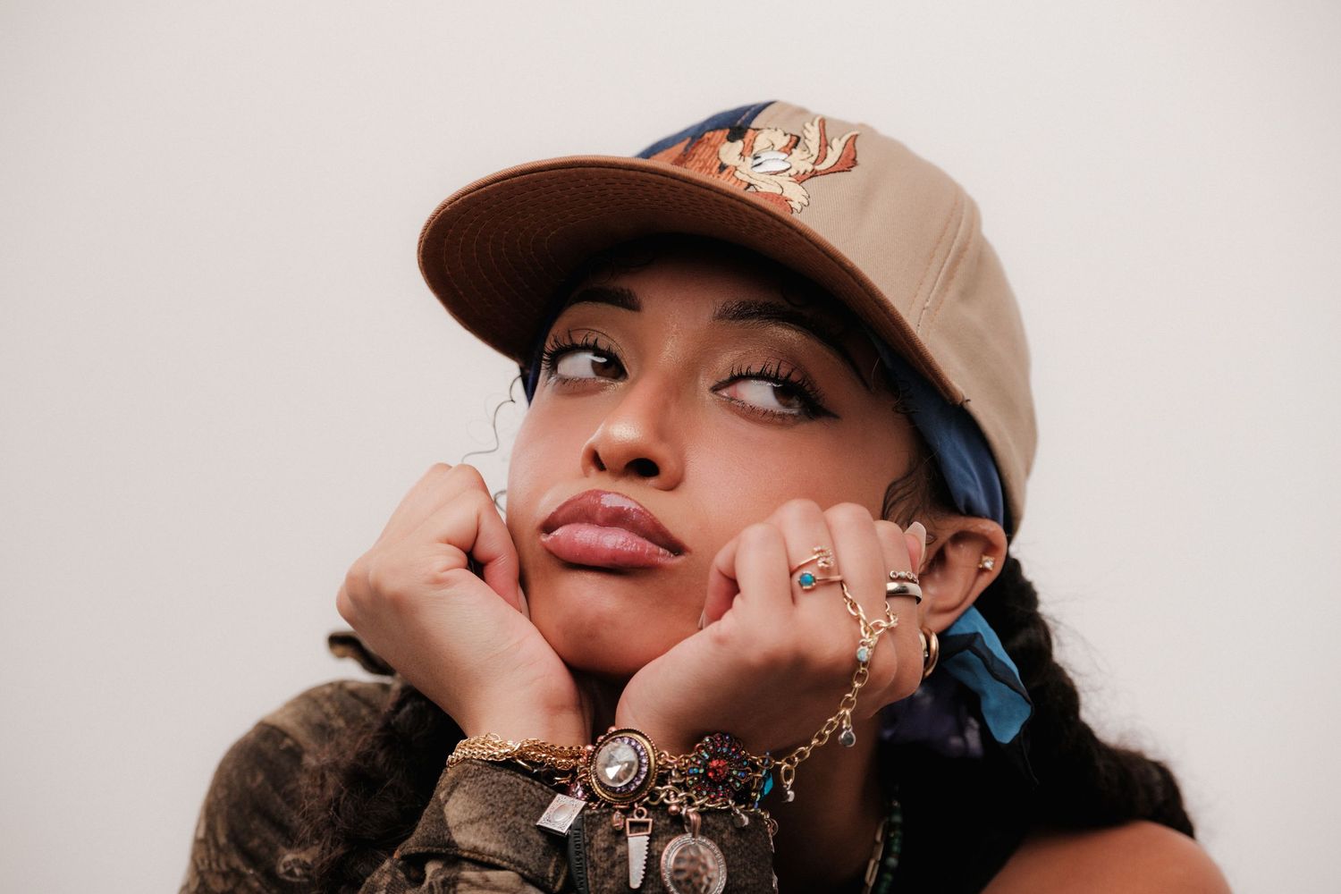 Young woman in cap and layered jewelry poses thoughtfully against light background.