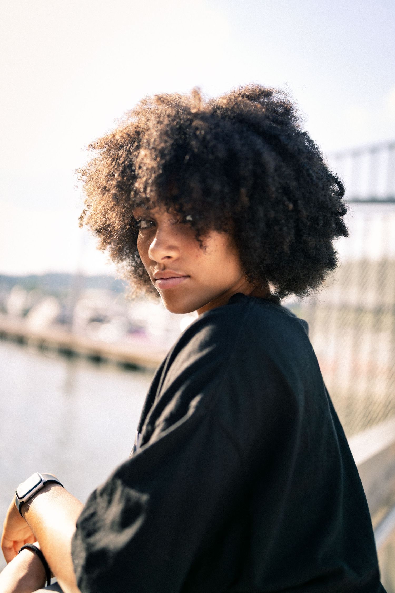 Young Black woman with natural afro hair poses outdoors near waterfront on sunny day.
