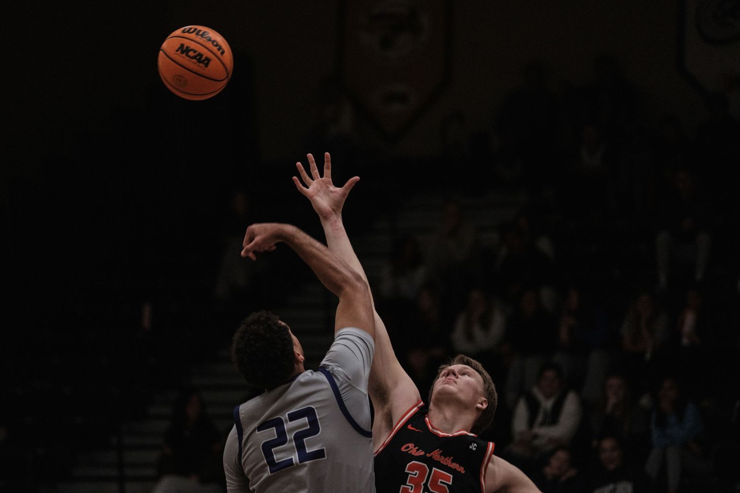 Two basketball players jumping for a tip-off during a competitive indoor basketball game.