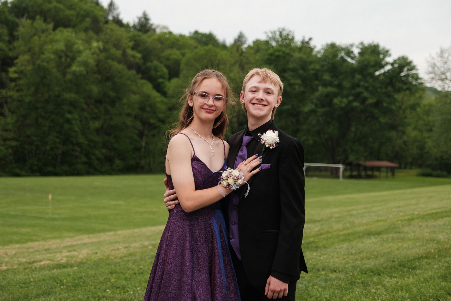 Teen couple in formal prom attire smile together on lush green field.