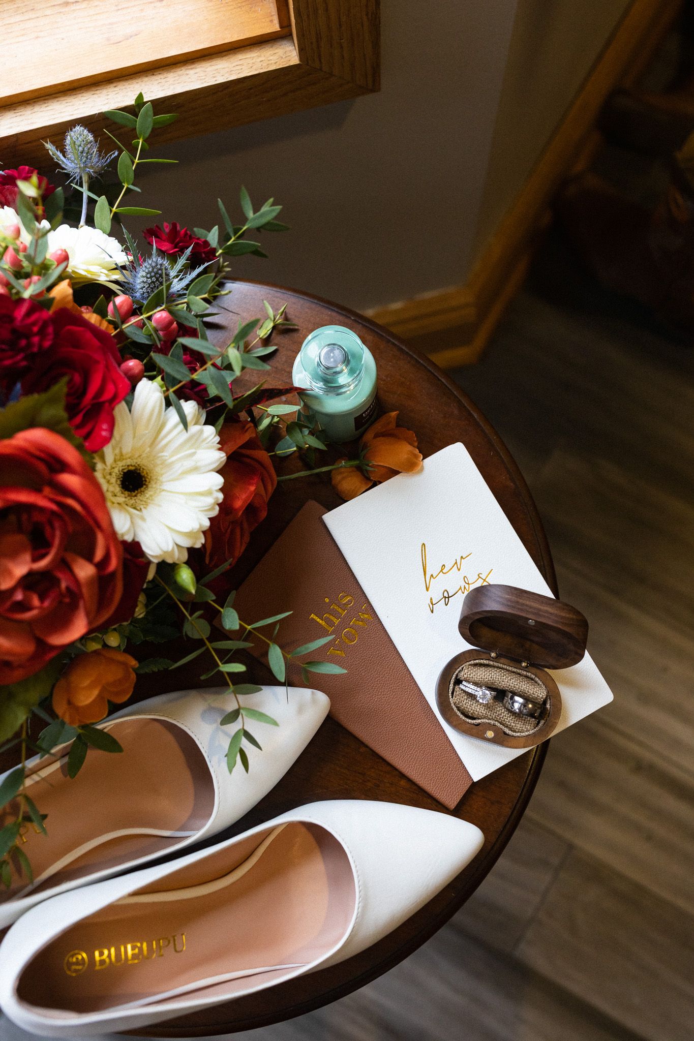 Wedding flat lay featuring white bridal heels, bouquet of red and orange flowers, rings, and vow book.