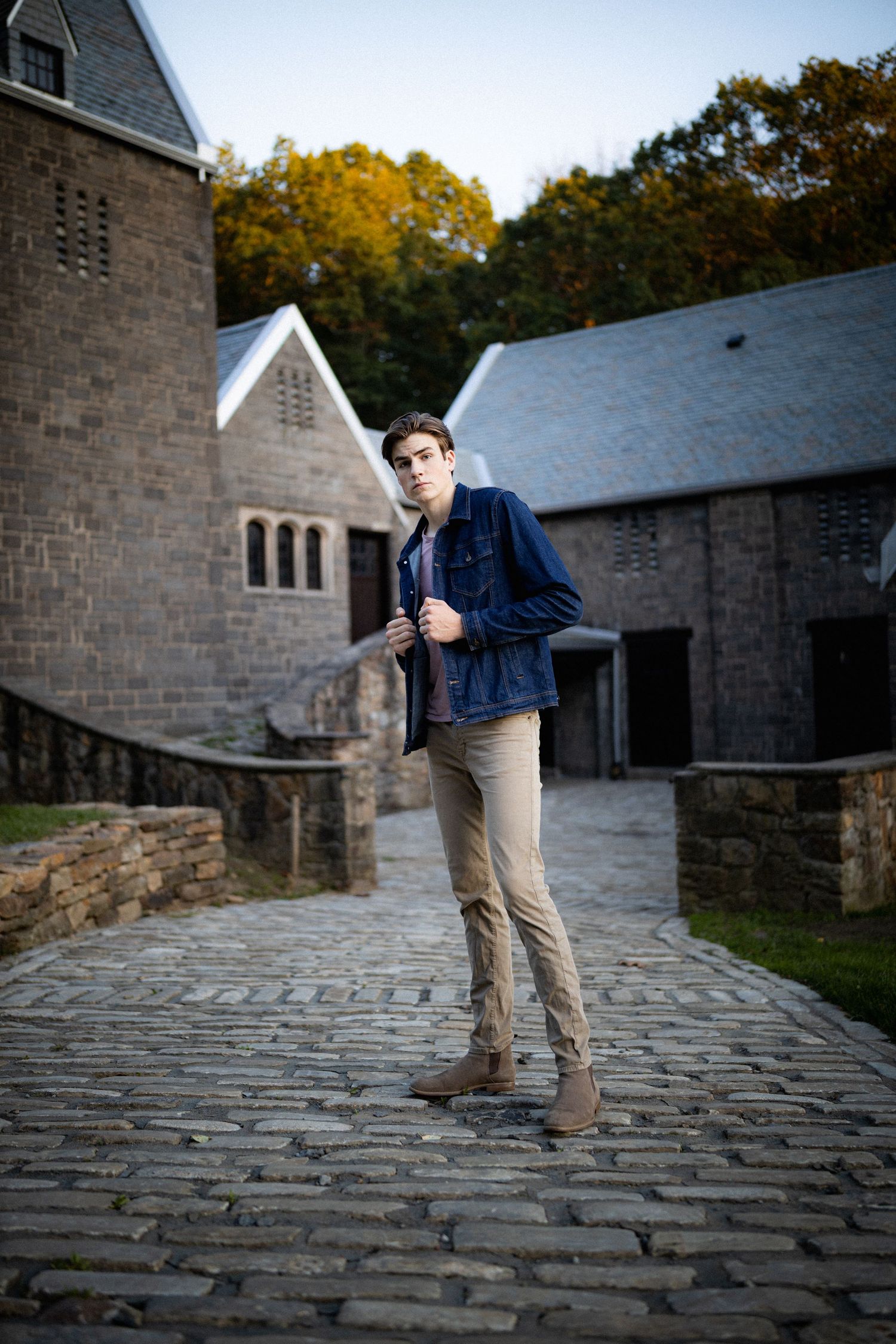 Stylish young man in denim jacket standing on cobblestone path near rustic stone buildings in autumn.