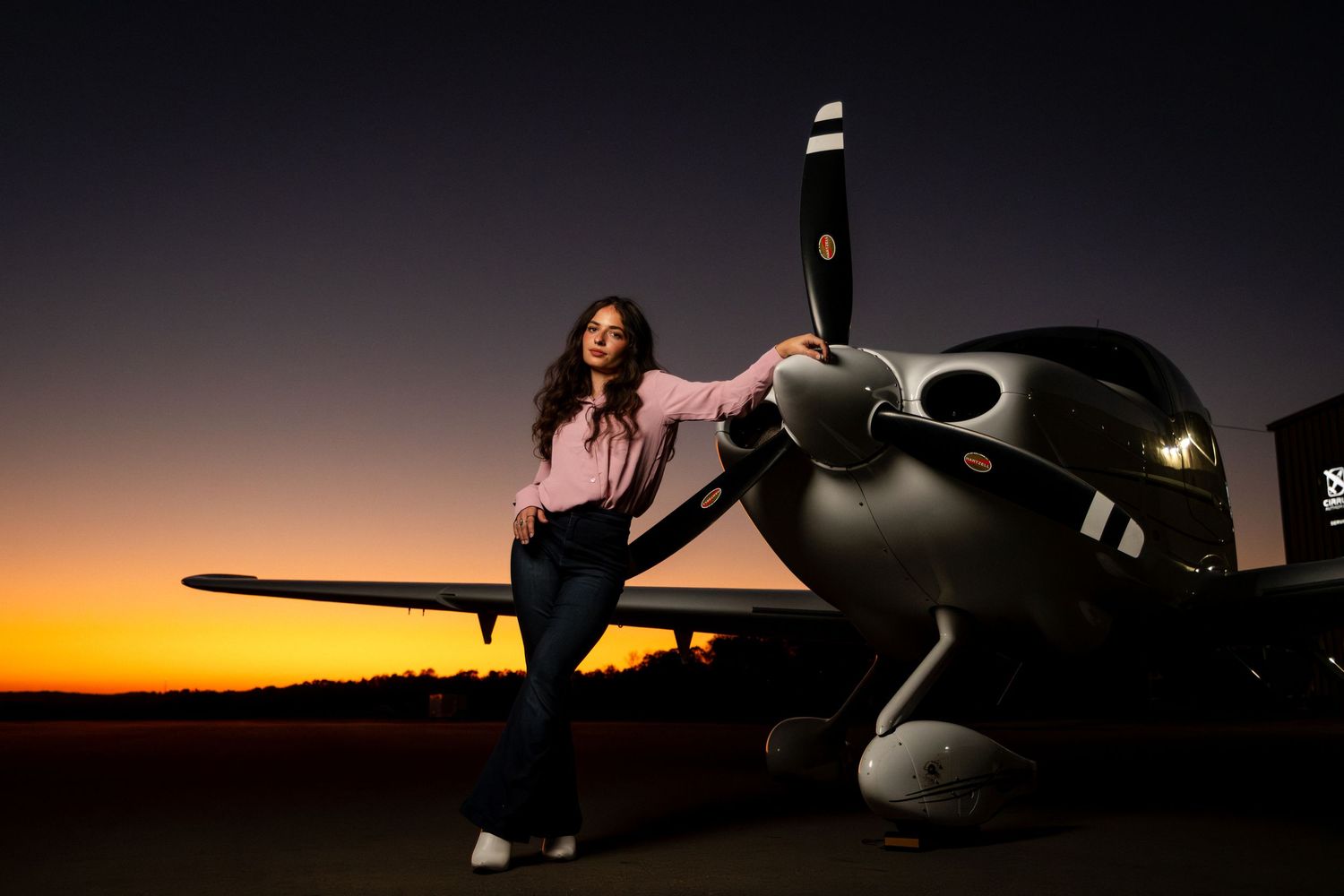 Young woman in pink shirt leaning against dark propeller aircraft at colorful sunset.