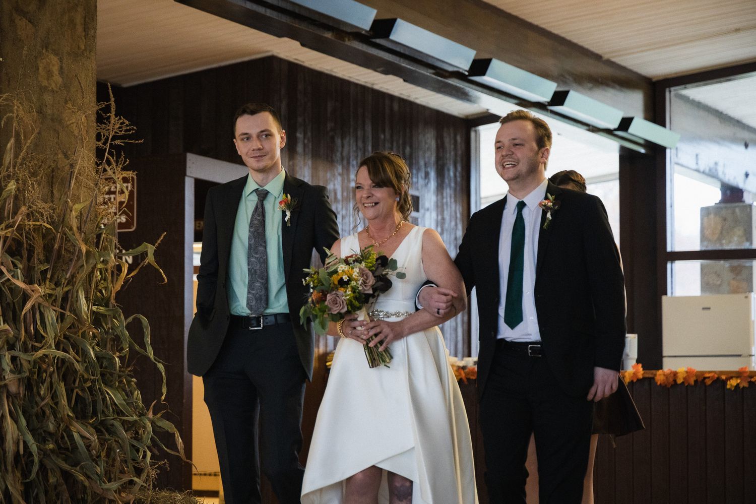 Bride holding bouquet flanked by two groomsmen in a modern wedding venue with autumn decor.