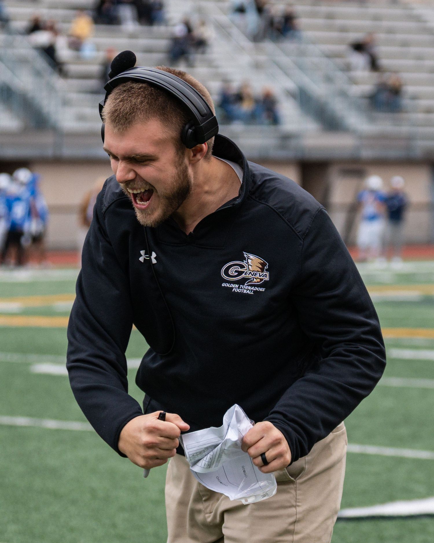 Excited Geneva College football coach in black jacket celebrates enthusiastically on sideline.