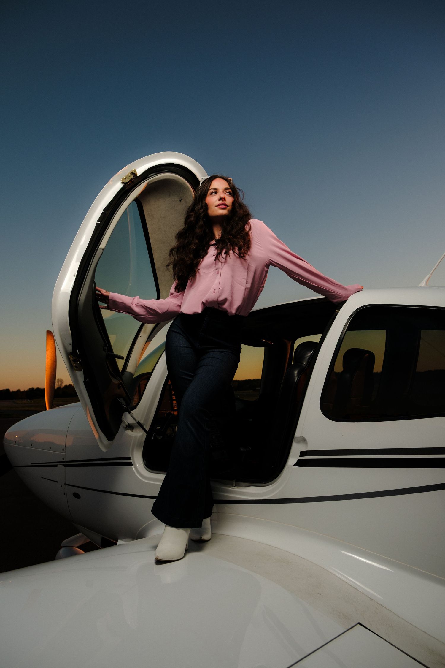 Young woman in pink blouse exiting small white propeller plane door at dusk.