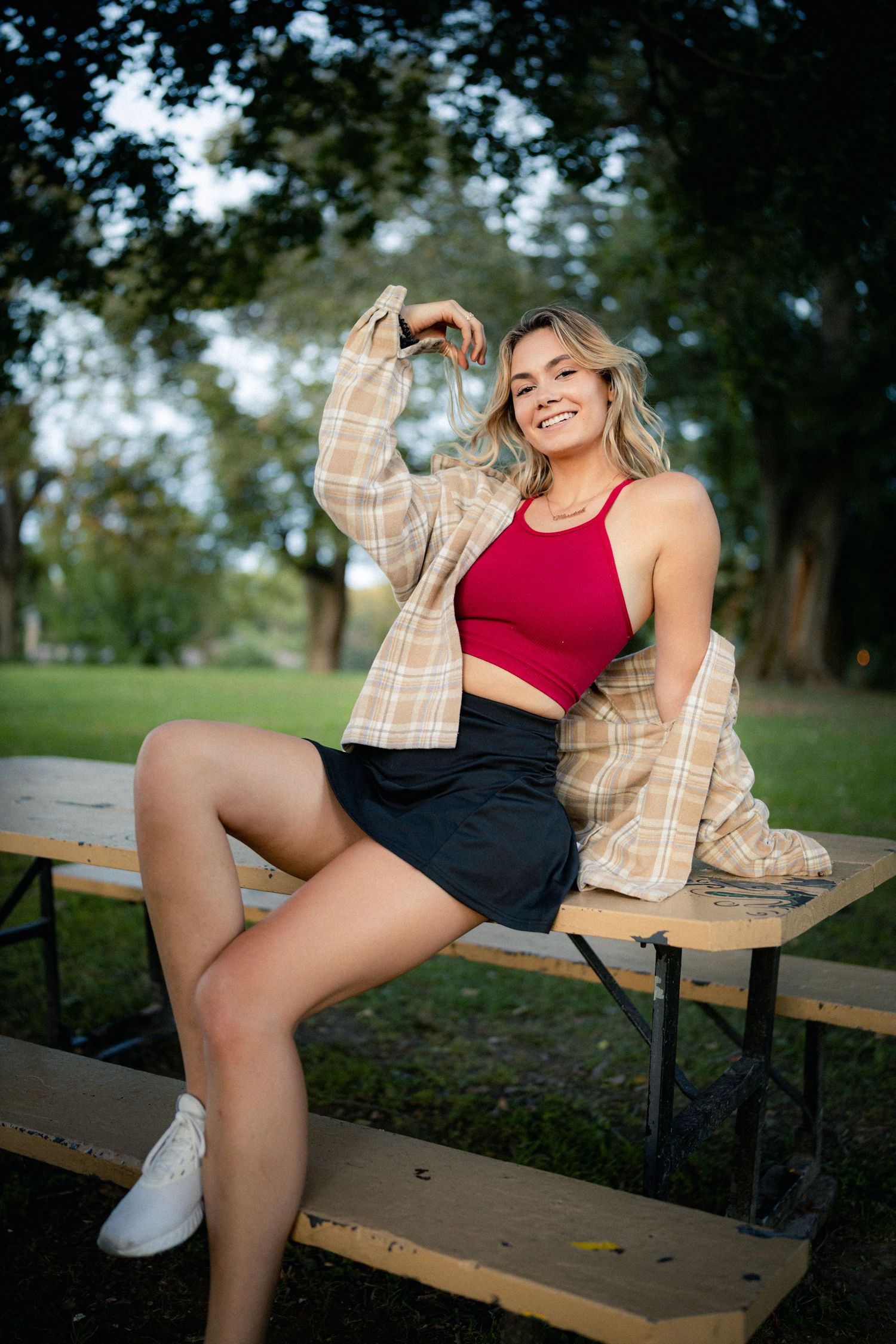 Cheerful young woman in red crop top and plaid shirt sitting on picnic table in green park.