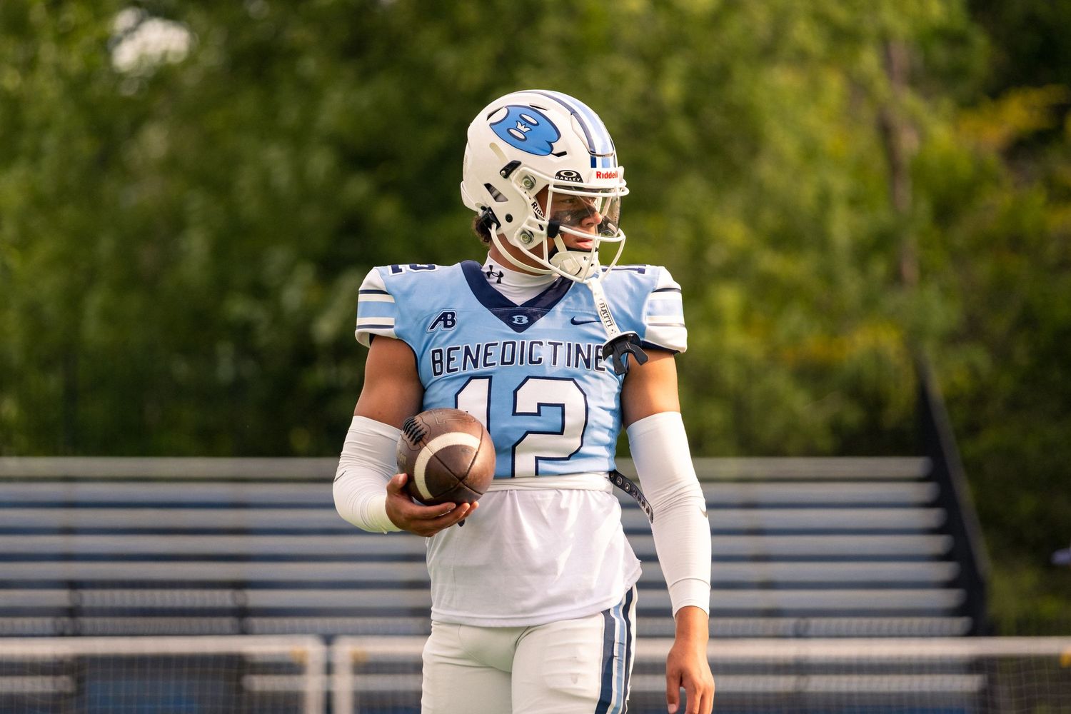 Benedictine football player number 12 in light blue uniform holds ball on sunny field.