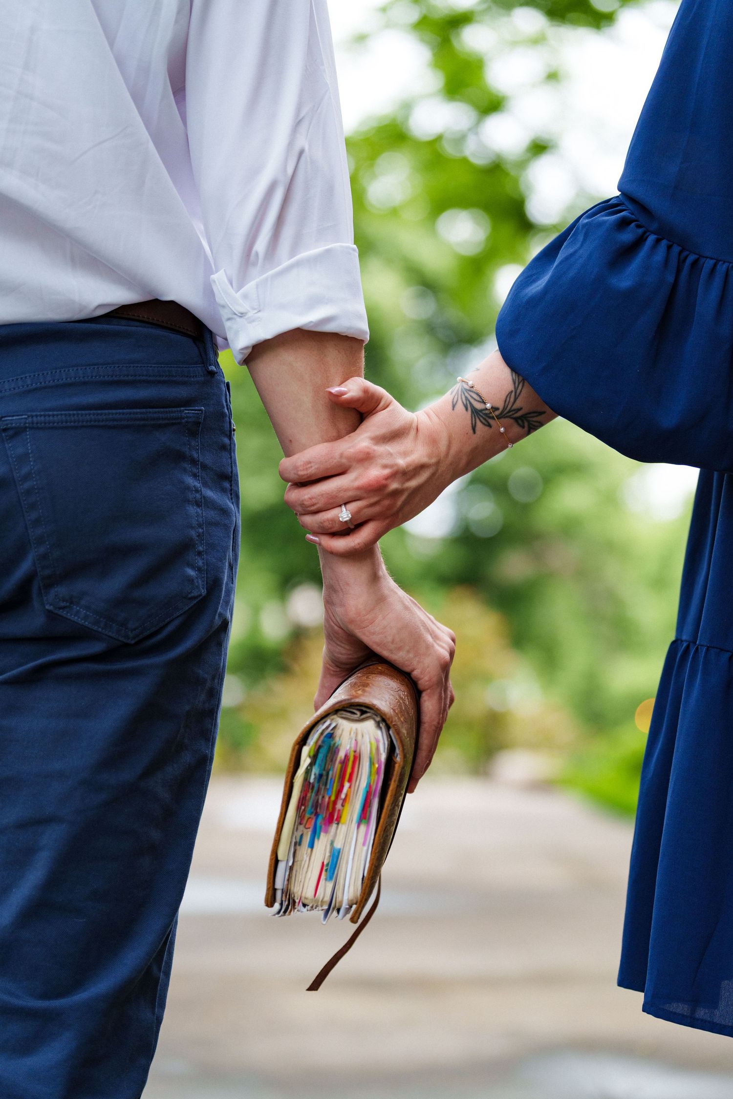 Close-up of a couple holding hands outdoors, one partner holding a colorful journal with a tattoo visible.