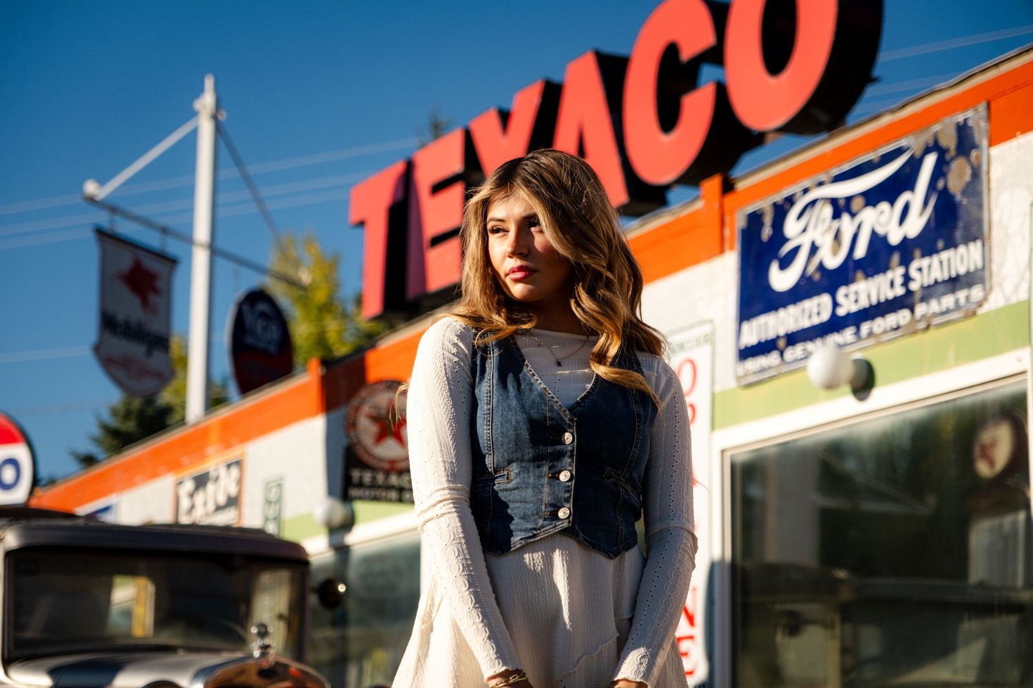 Young woman in denim vest poses before vintage Texaco gas station on sunny day.