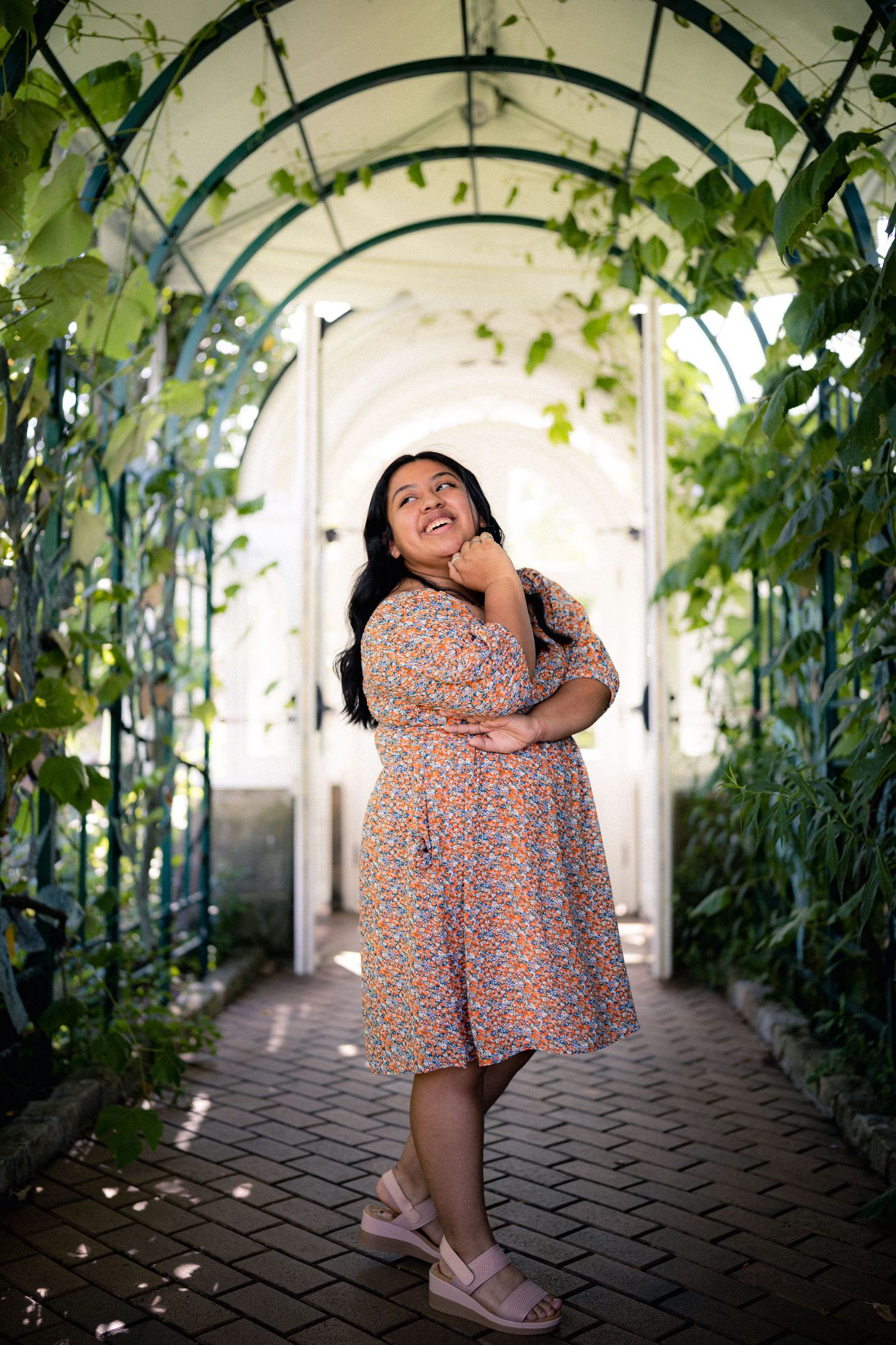 Young woman in floral dress smiling under arched garden tunnel with lush green vines.