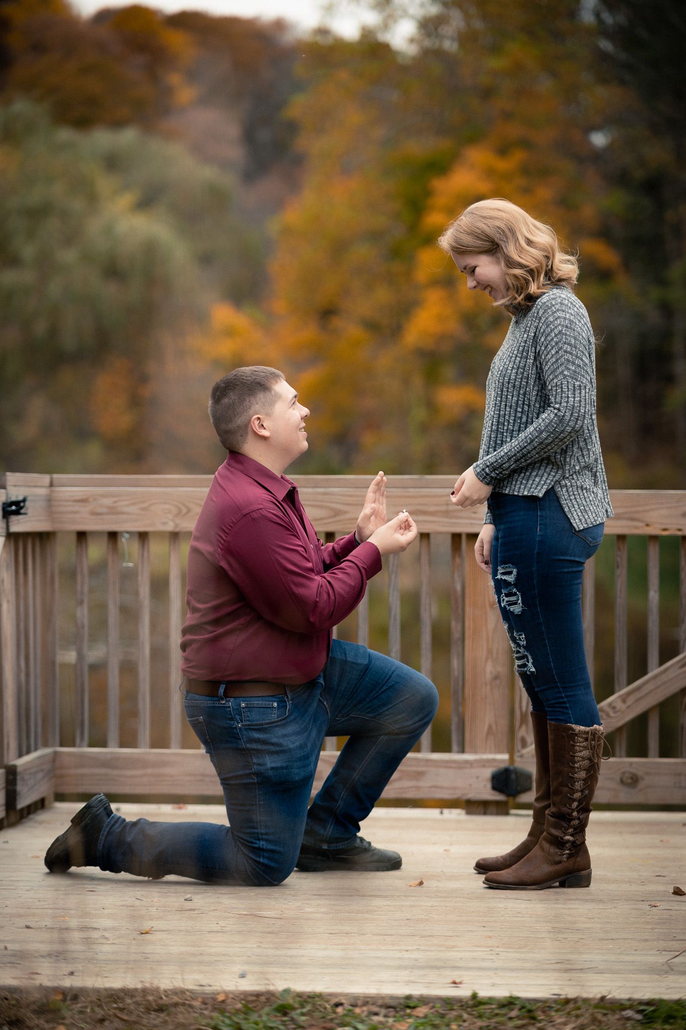 Man proposing on one knee to his girlfriend on a wooden bridge surrounded by fall foliage.