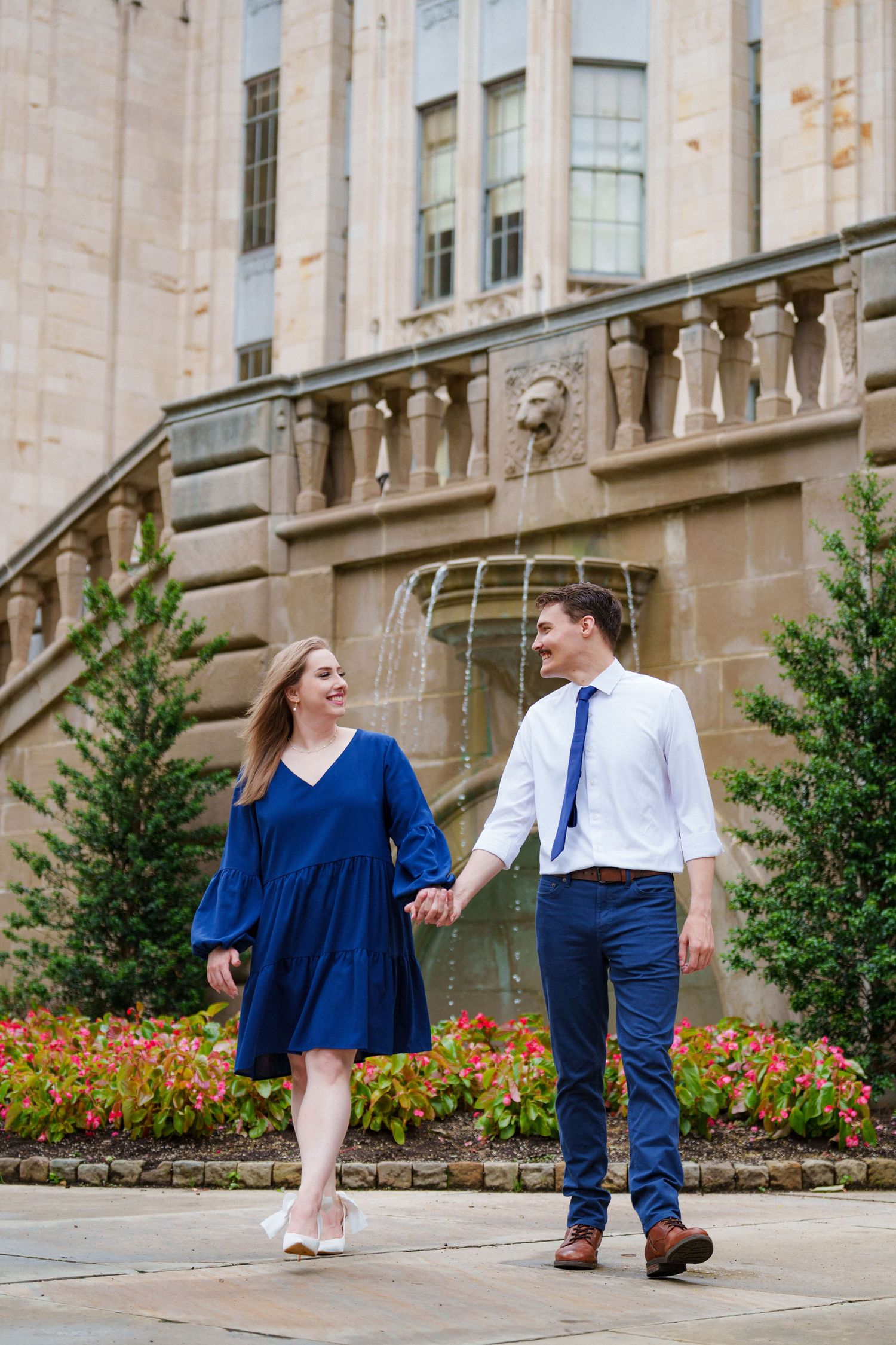 Smiling couple walking hand-in-hand near a fountain outside a grand stone building with colorful flowers.