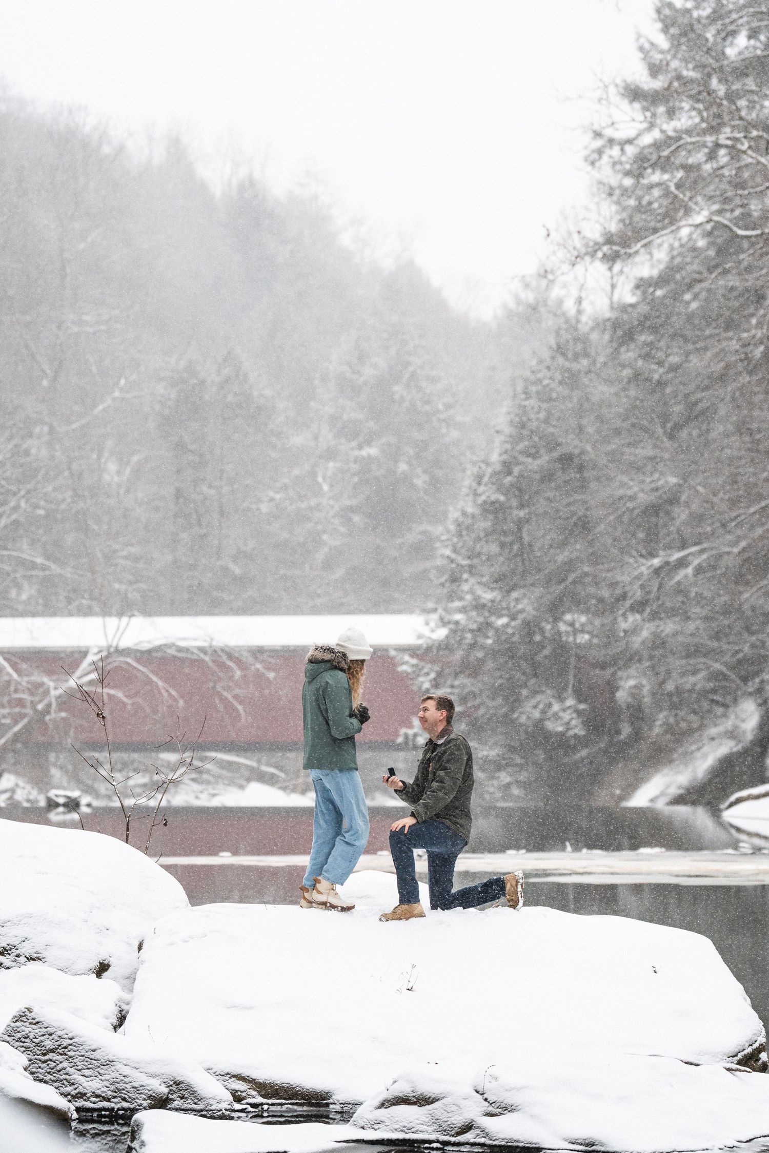 Man proposes on one knee in a snowy winter landscape near a red covered bridge during a snowstorm.