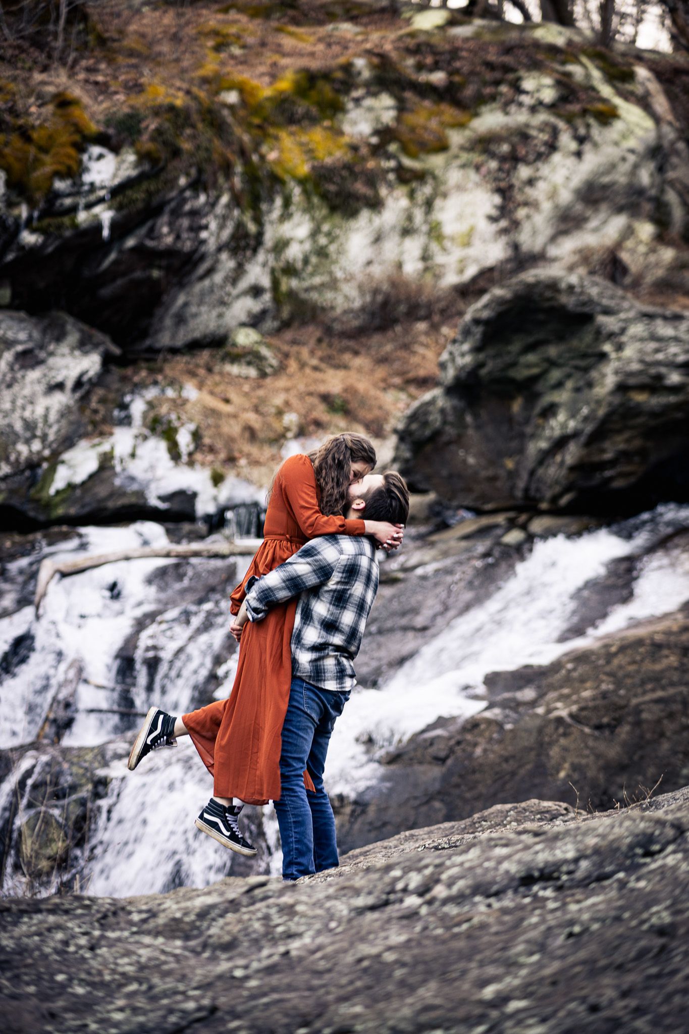Couple sharing a lift and kiss near rocky waterfall, woman in rust dress and man in plaid shirt.