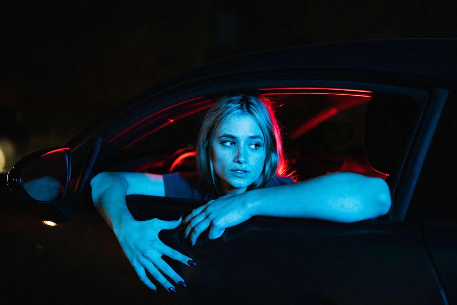 Blonde woman leans out of car window bathed in blue and red neon lights at night.