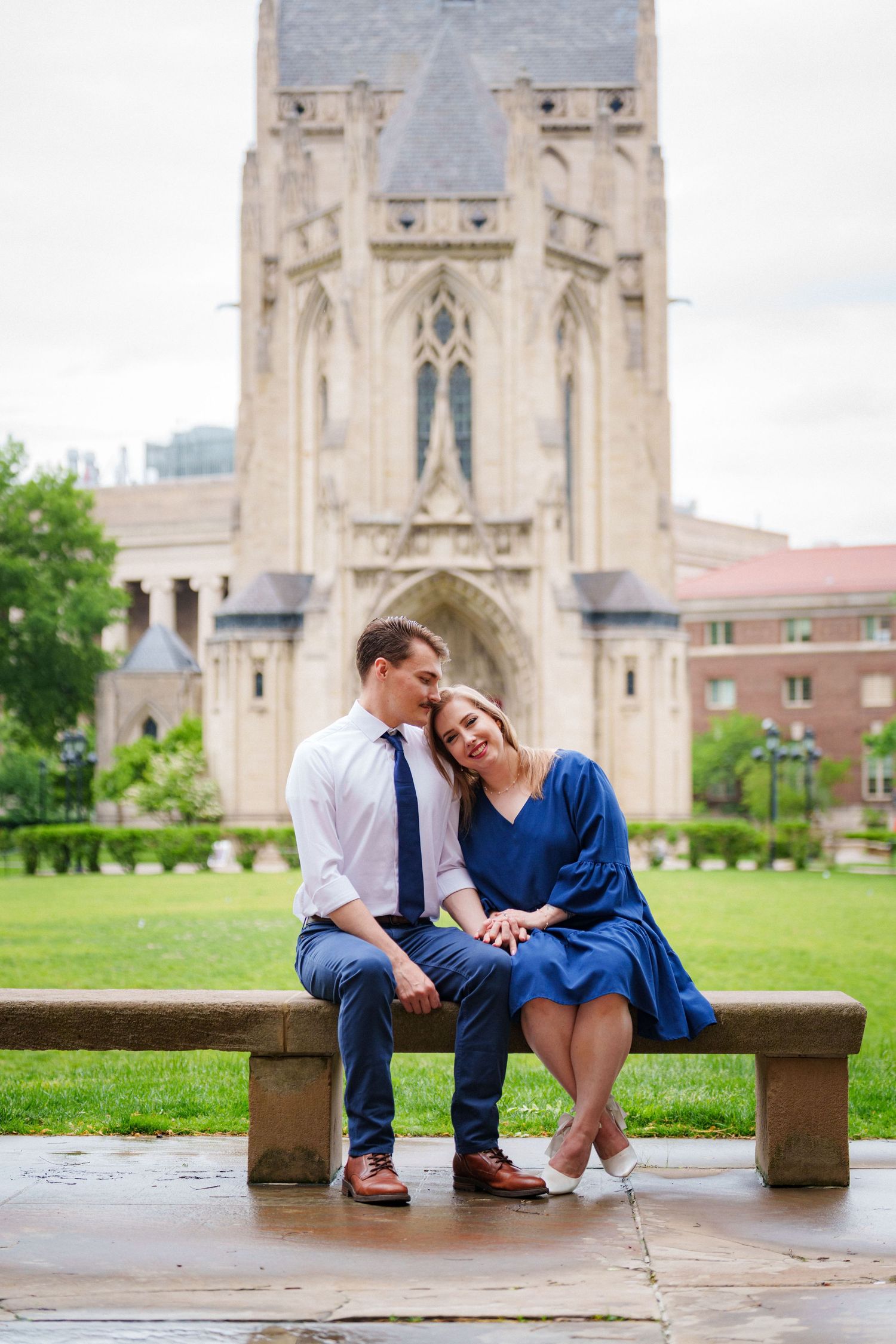 Romantic couple sitting closely on a stone bench in front of a stunning Gothic cathedral on a cloudy day.