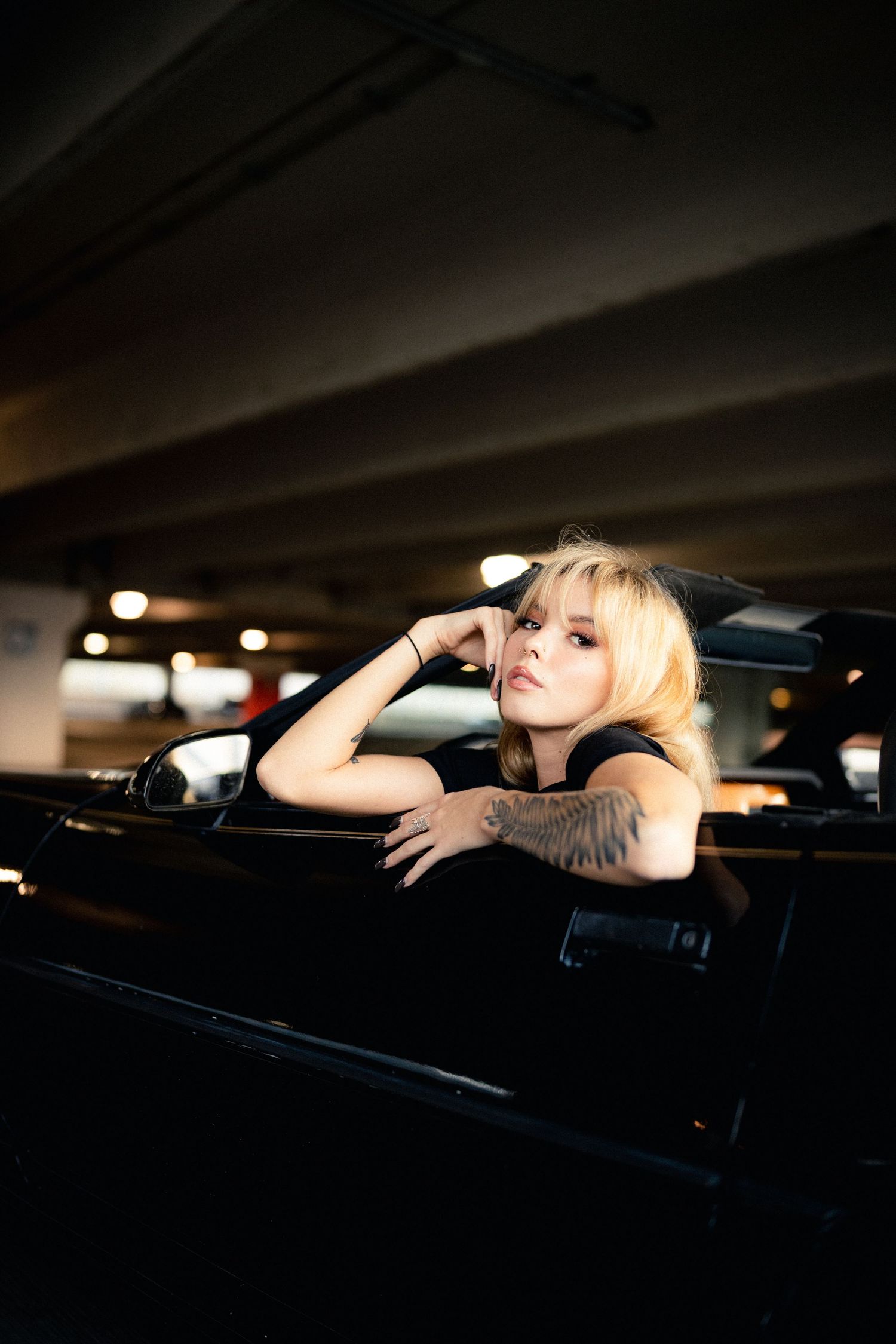 Edgy blonde woman with tattoos leaning out of black car window in dimly lit parking garage.