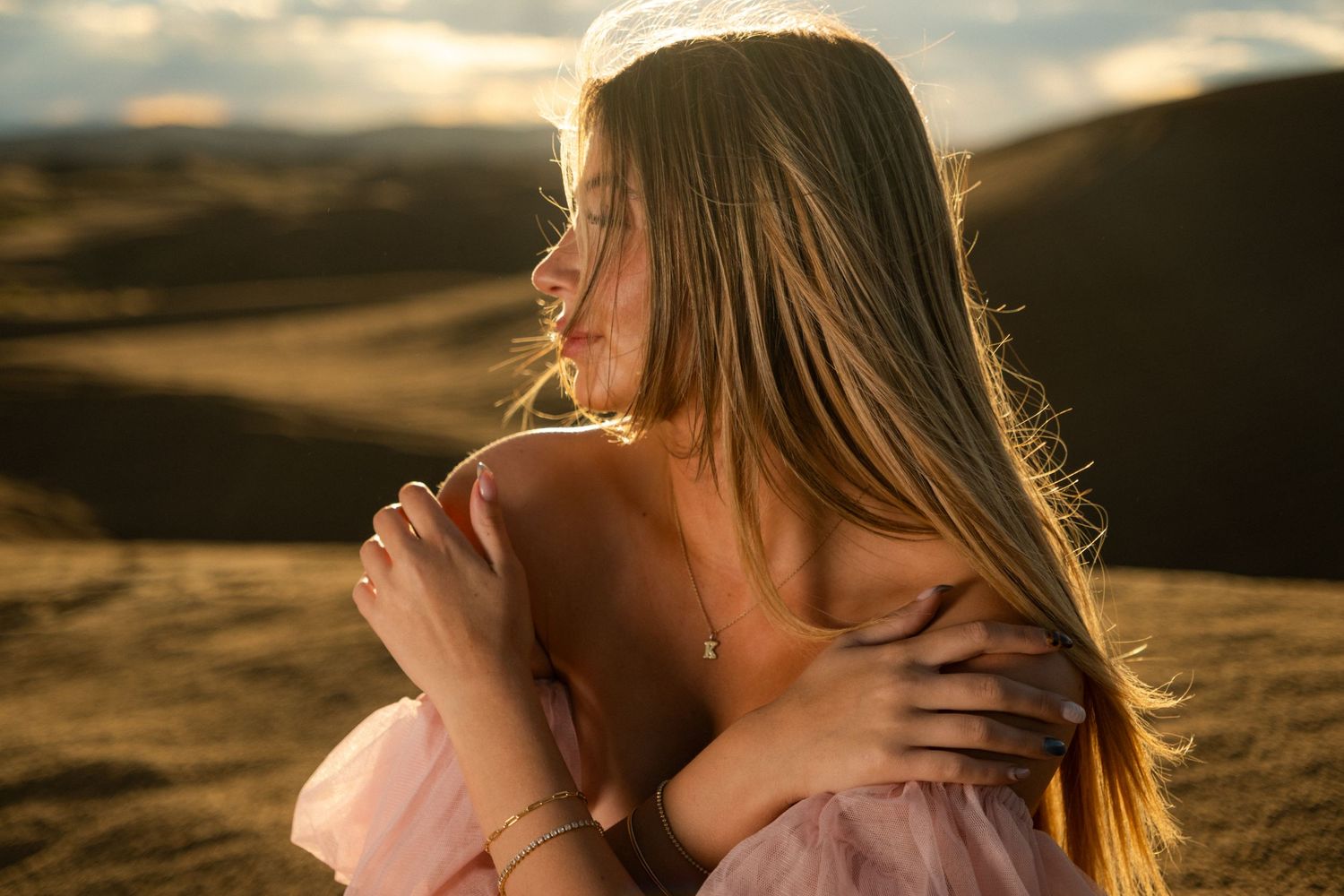 Young woman in pink dress hugs knees while sitting in golden sunlit field.