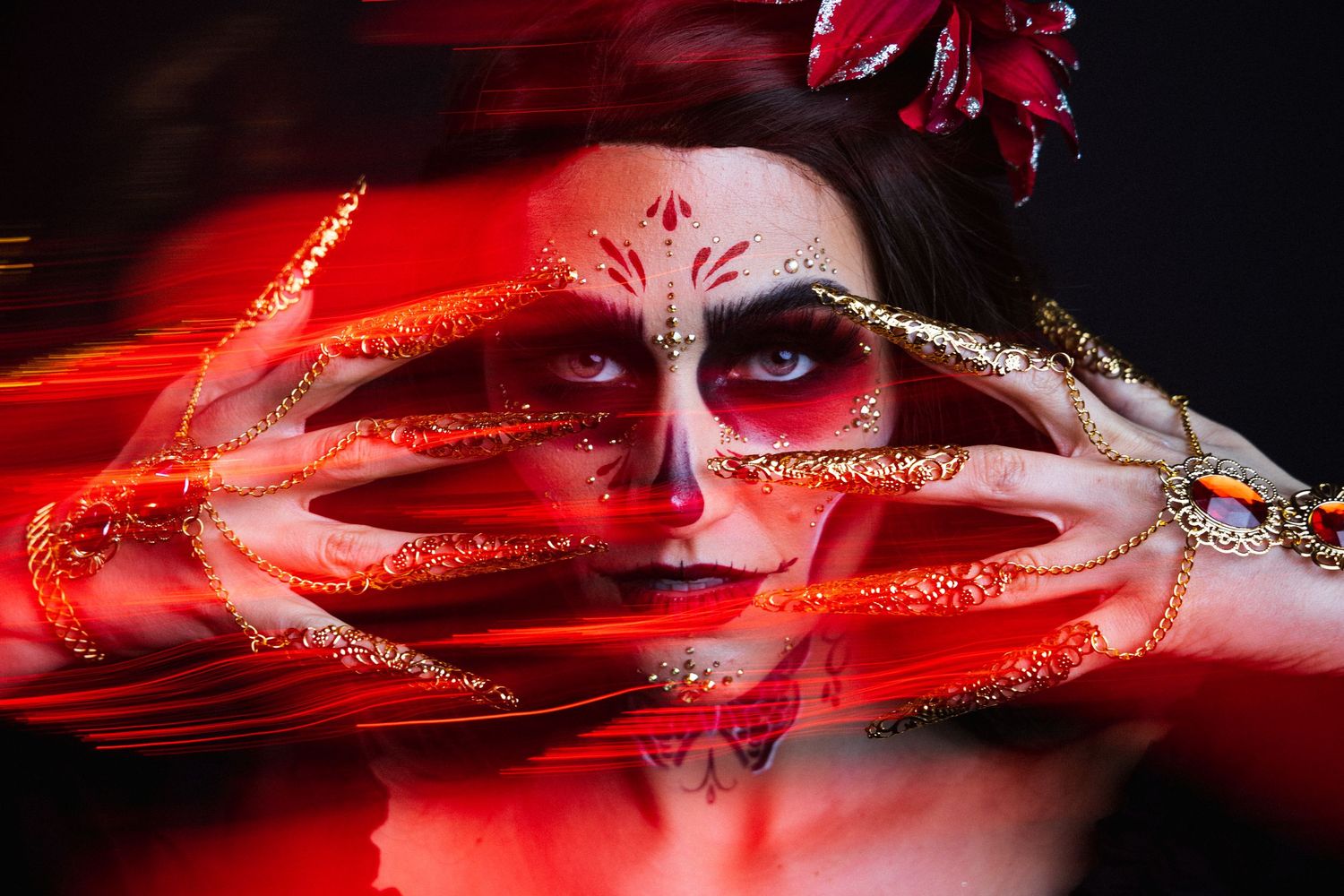 Woman with elaborate Day of the Dead makeup and gold nails surrounded by red light streaks.